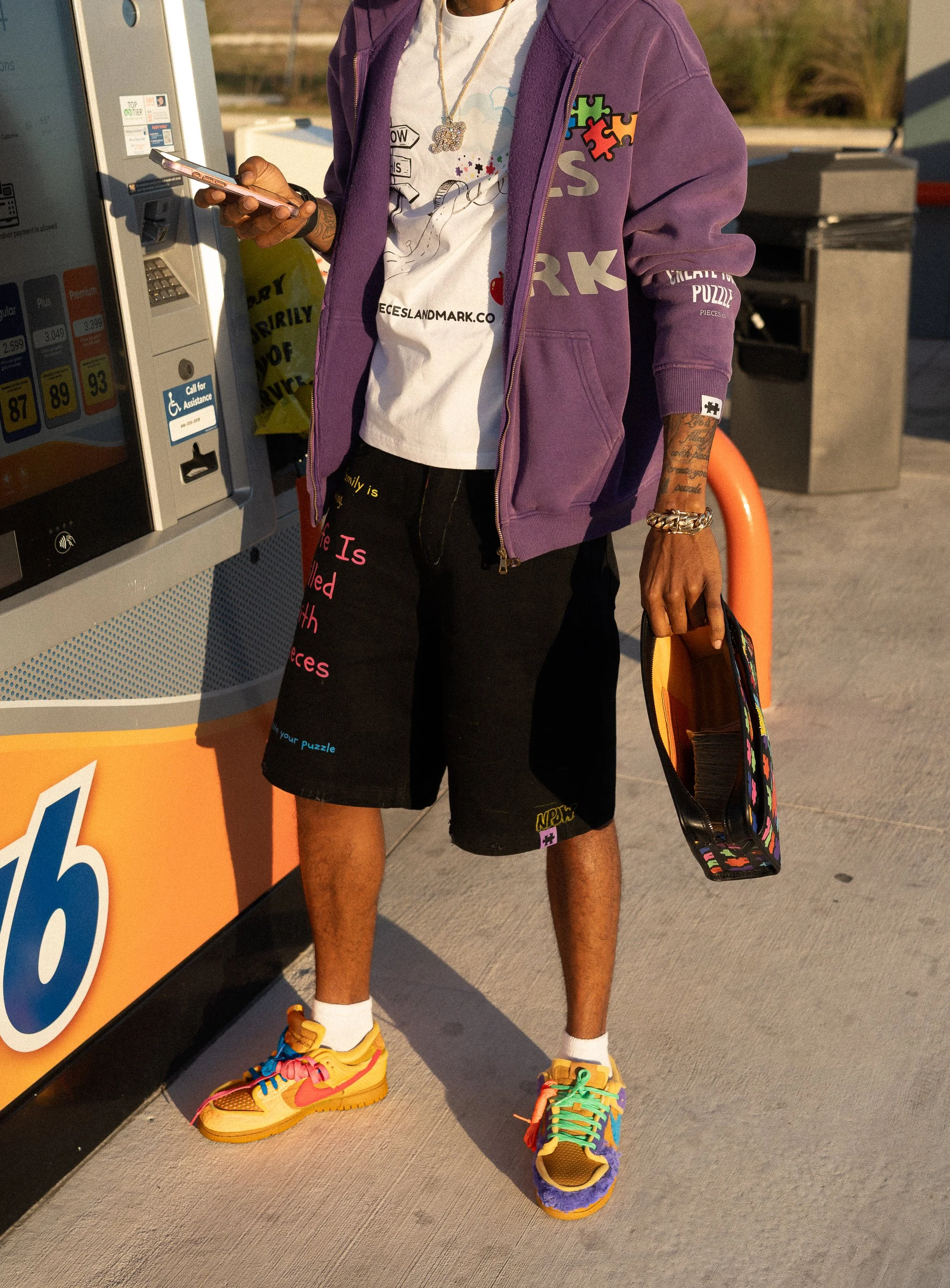A person standing at a vending machine, wearing colorful sneakers, black shorts, a white printed T-shirt, a purple hoodie, and various jewelry, holding a phone and a bag.