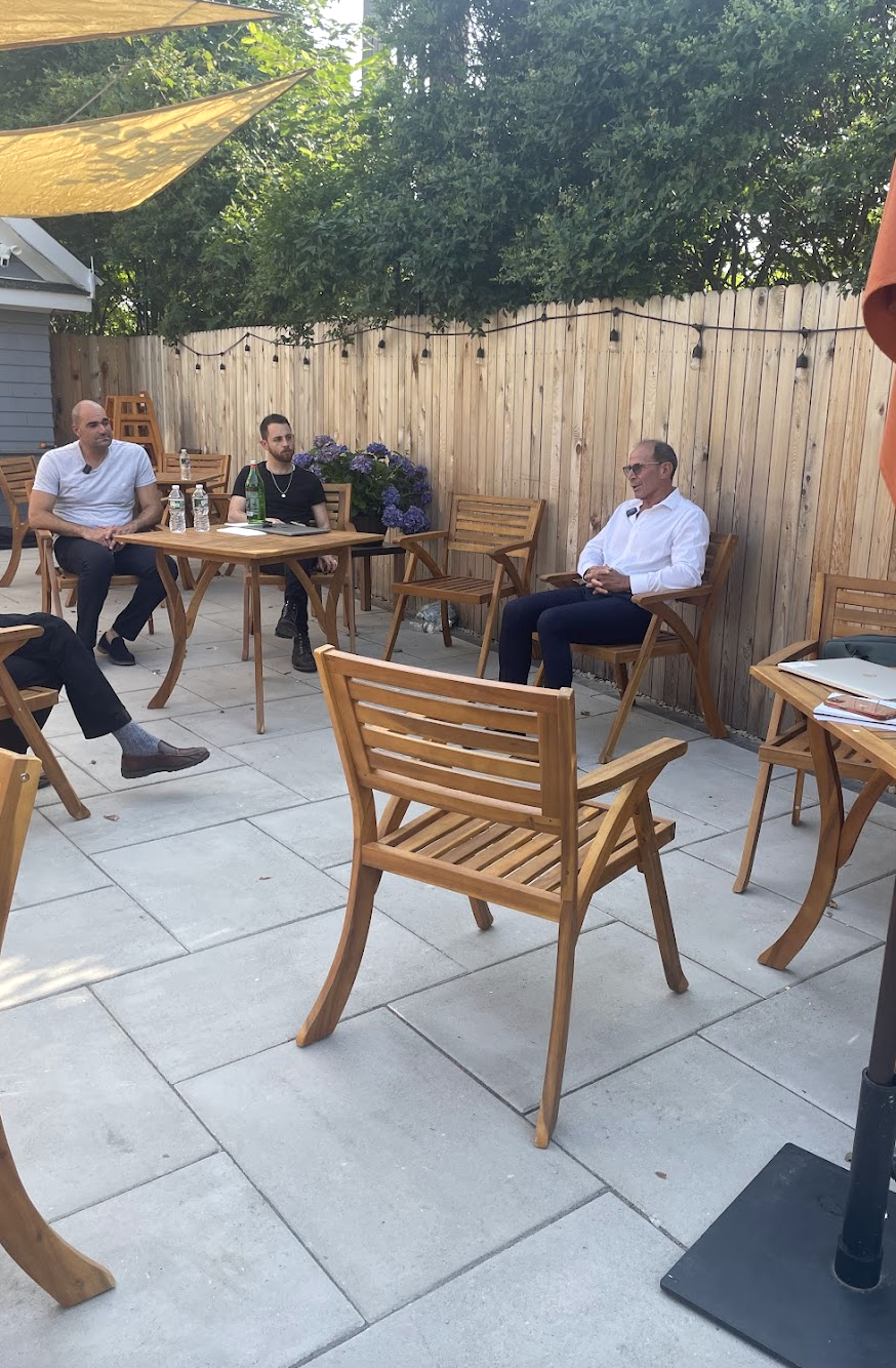 Three men are sitting and listening in an outdoor patio with wooden chairs, a wooden fence, and string lights, with a woman partially visible on the right. There are water bottles on the tables and trees overhead.