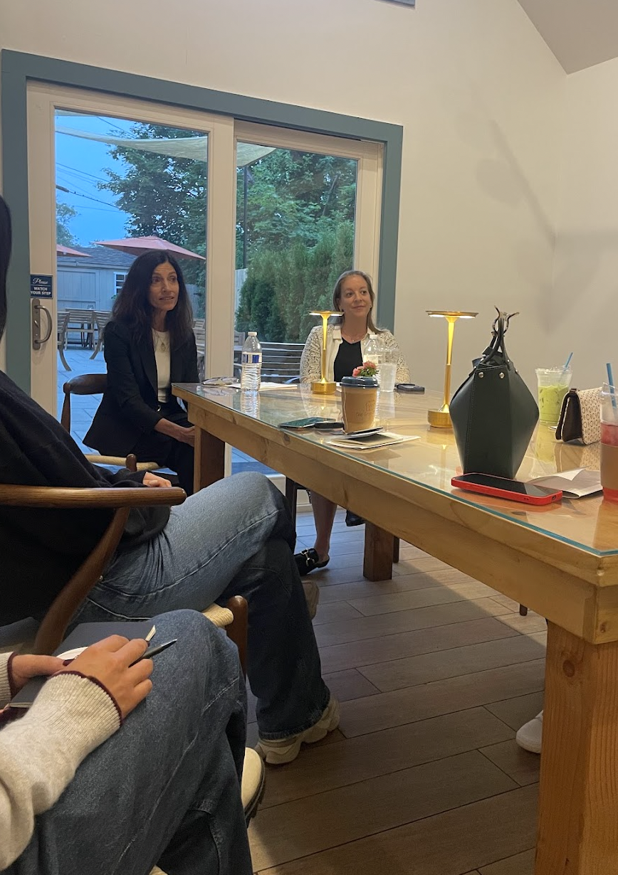 Women sitting around a wooden table in a room with large glass sliding doors showing an outdoor patio and trees.