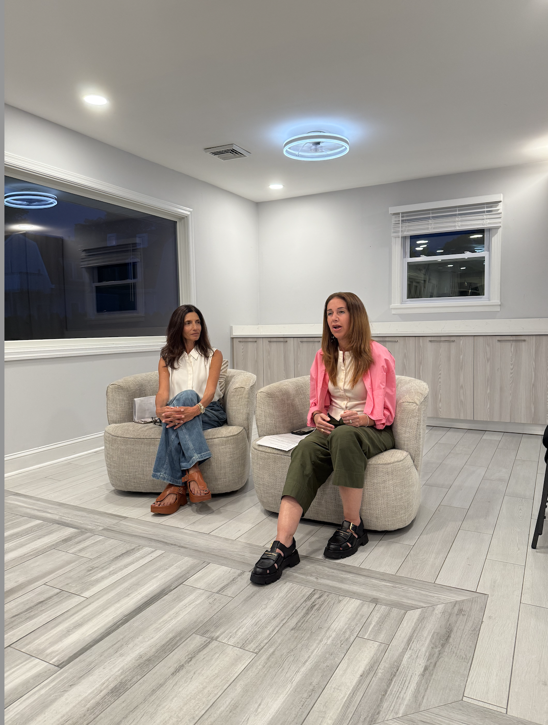 Two women sitting on beige chairs in a modern, minimally decorated room with white walls, large windows, and hardwood floors, engaged in conversation.