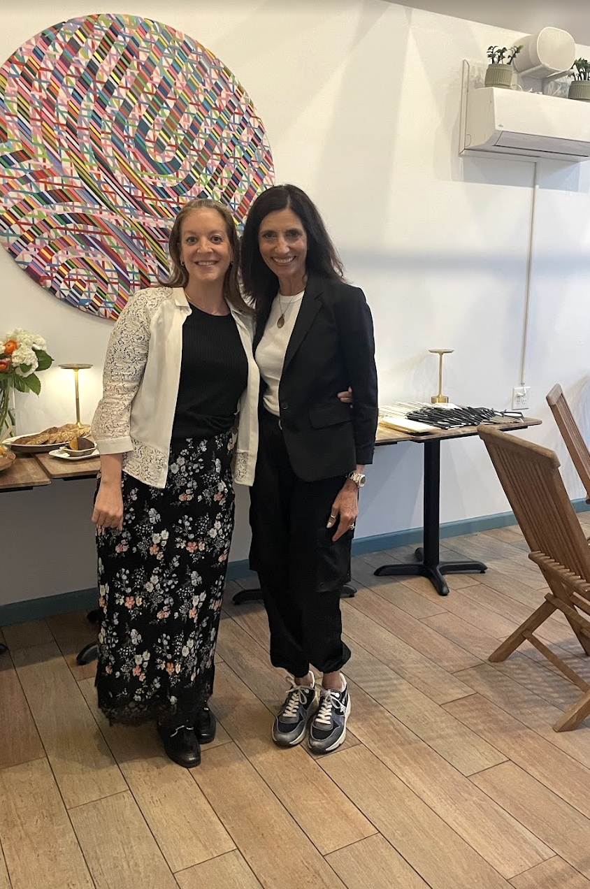 Two women standing side by side in a room with a colorful circular artwork on the wall behind them. One woman is wearing a white lace cardigan over a black top and a floral skirt, while the other is dressed in a black blazer, white shirt, black pants