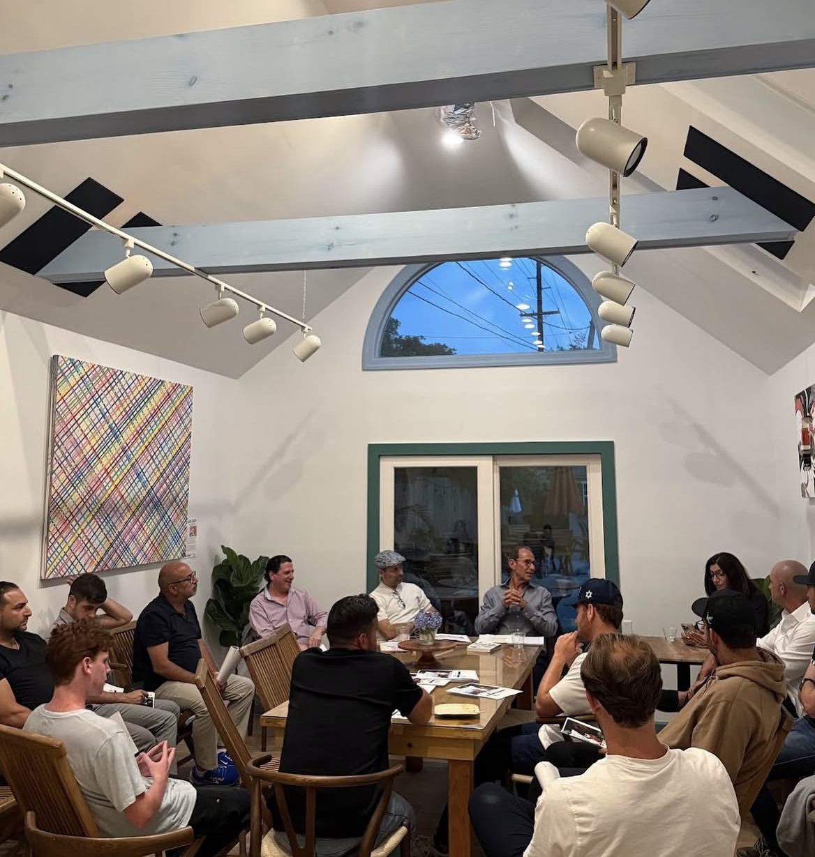 People seated at a meeting in a modern room with a high, vaulted ceiling, large window, and abstract artwork on the wall.