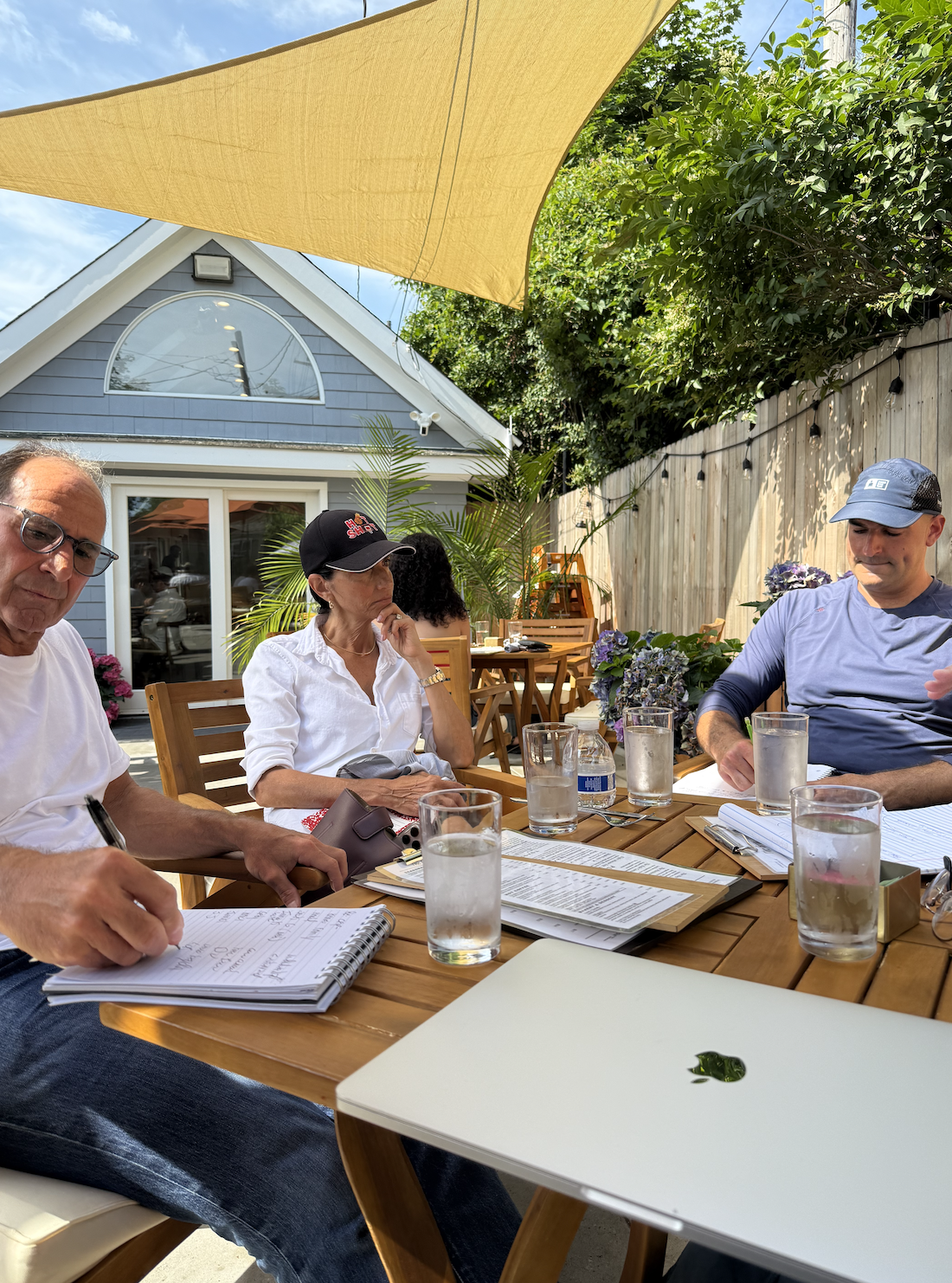 People sitting at a wooden outdoor table under a large yellow shade sail, with a house and greenery in the background. They are engaged in a discussion, with notebooks, pens, and glasses of water on the table.