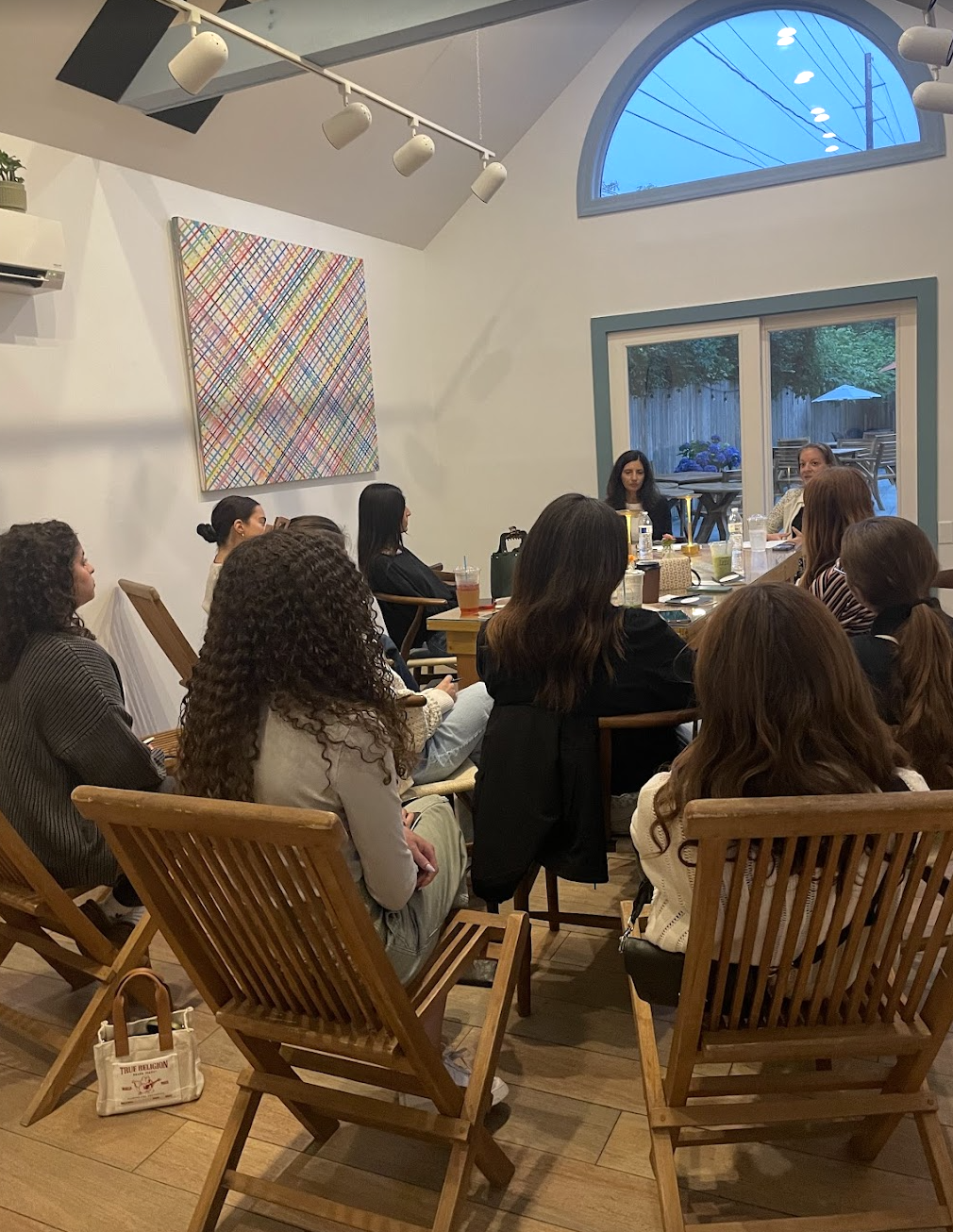 A group of women seated around a rectangular table in a modern dining room, listening to a speaker at the head of the table. The room has white walls, a colorful abstract painting on one wall, and large windows showing an outdoor patio with a table a