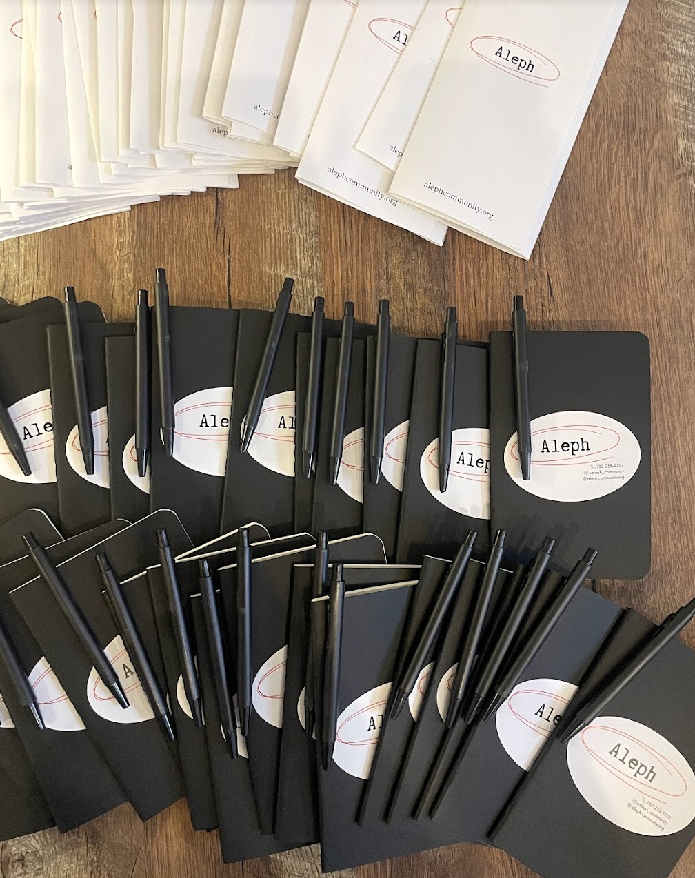 Multiple black notebooks with pens and stickers labeled 'Aleph' on a wooden table.