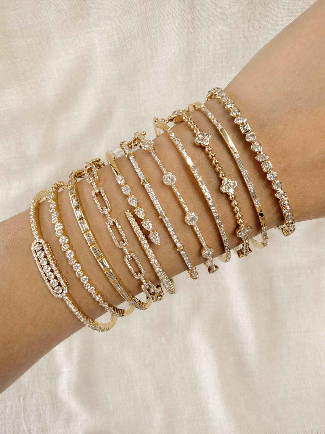 A person's arm adorned with multiple gold and silver bracelets featuring various designs and embedded with diamonds, displayed against a beige fabric background.