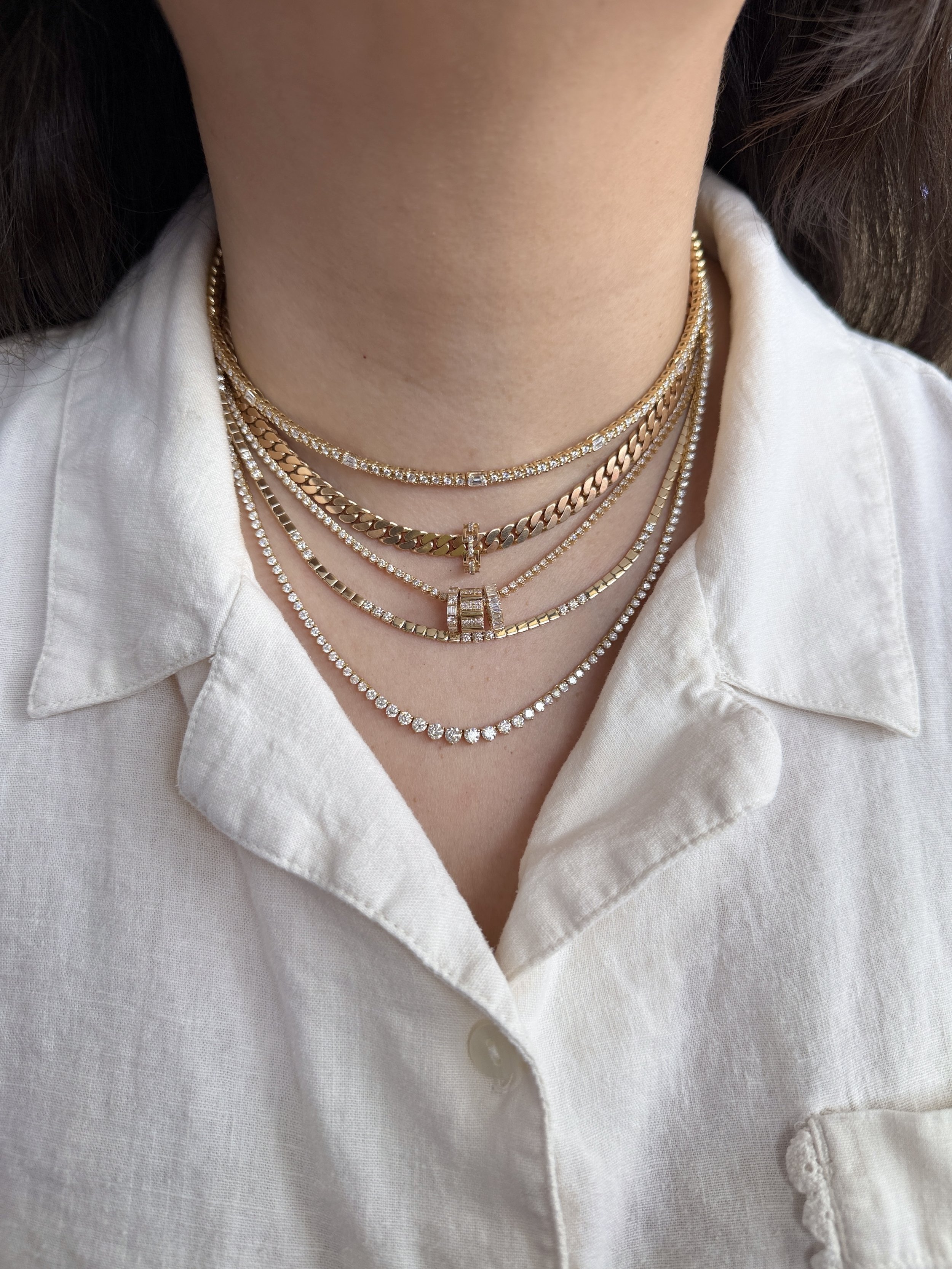 Close-up of a person's neck and chest, adorned with multiple layered gold and diamond necklaces, wearing a white collared shirt.