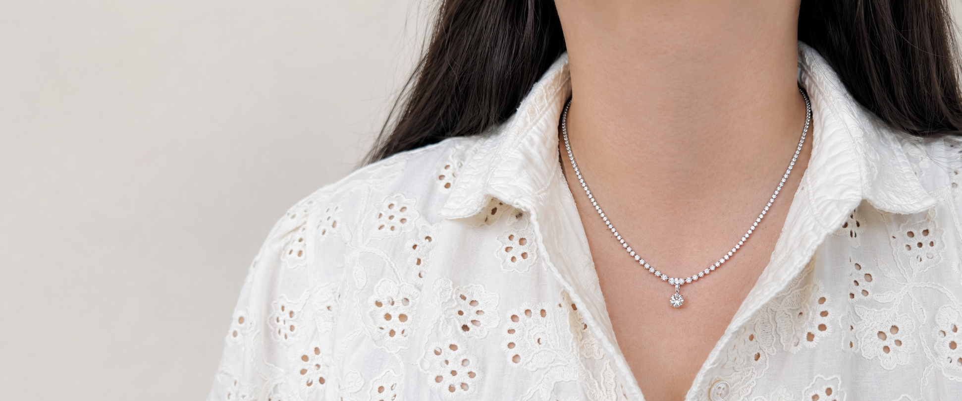 Close-up of a woman wearing a white embroidered blouse with eyelet details, a diamond necklace with a pendant, and long dark hair.