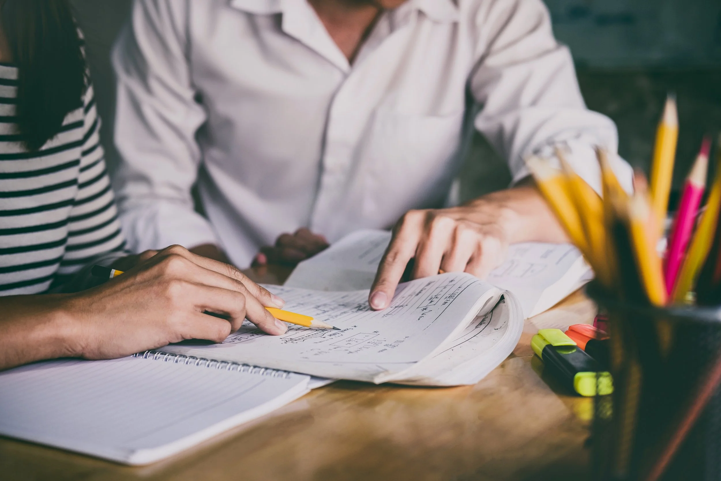 Two people are working on a project together at a desk with notebooks, highlighted pens, and colorful highlighters. This photo illustrates tutoring provided by The Margaret "Peggy" Krachy Children's Charity (aka the Peggy Principle)