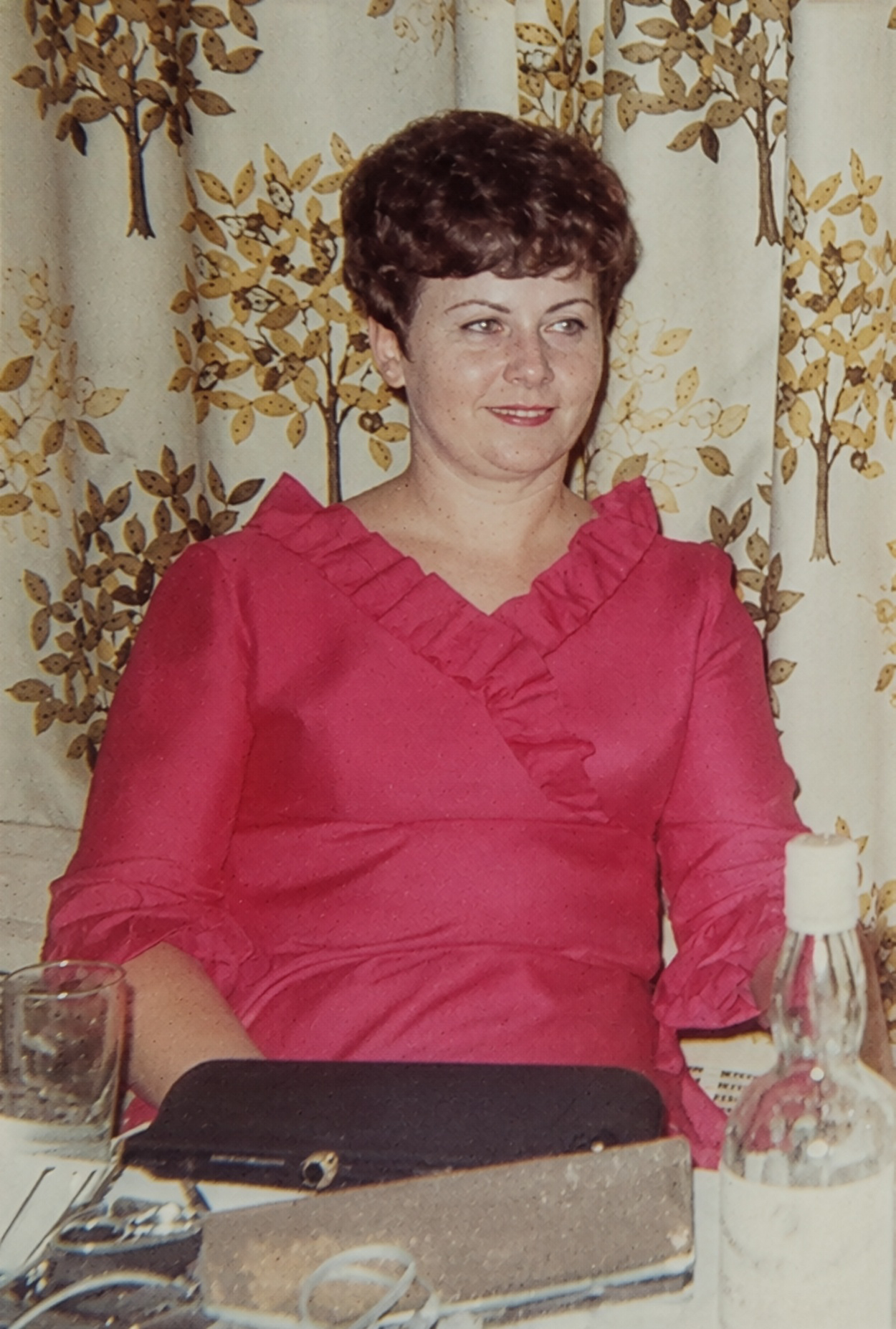 A photo of a young Peggy Krachy shown here wearing a pink dress with ruffled collar and sleeves, sitting at a table with a patterned curtain in the background, and a beverage bottle and glass in front of her.