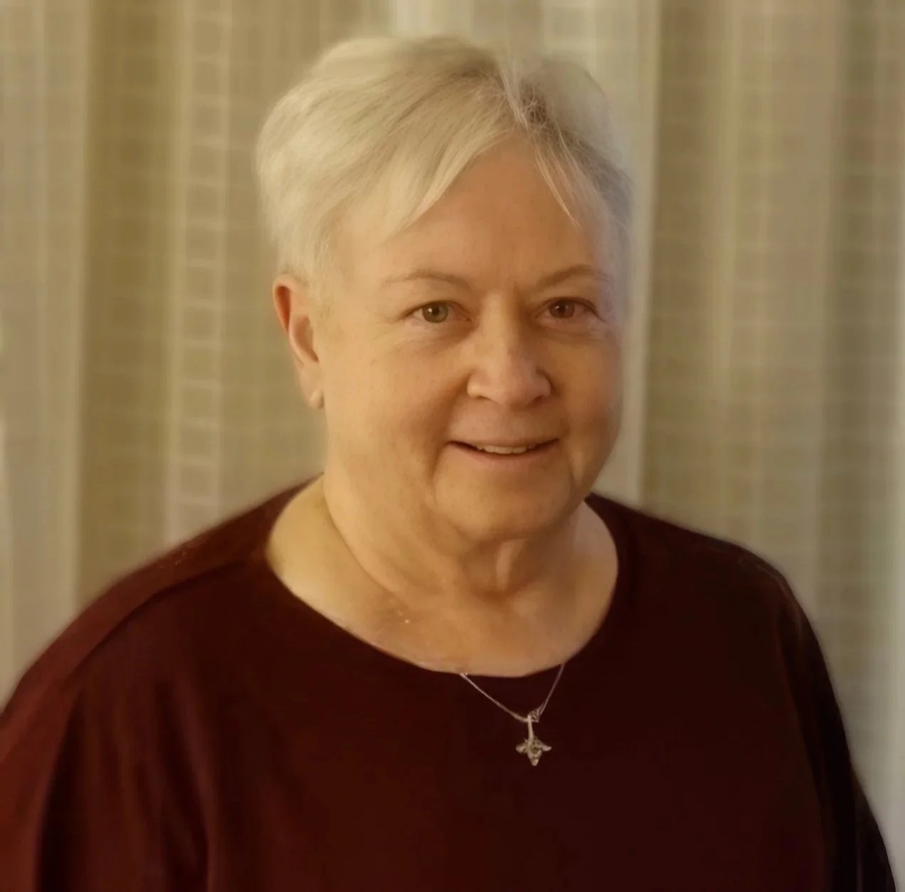 A photo of Peggy Krachy with short gray hair wearing a dark red top and a silver necklace with a cross pendant, smiling at the camera with a neutral curtain background.