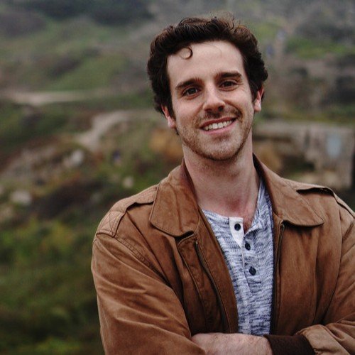 Portrait of a man smiling outdoors in a rural area with hills and greenery in the background.