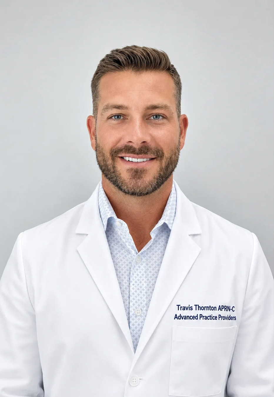 A smiling man in a white medical coat with text on it, standing against a plain background.