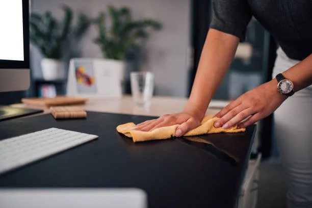 Person cleaning a dark office desk with a yellow cloth, with a computer monitor, glass, and plants in the background.