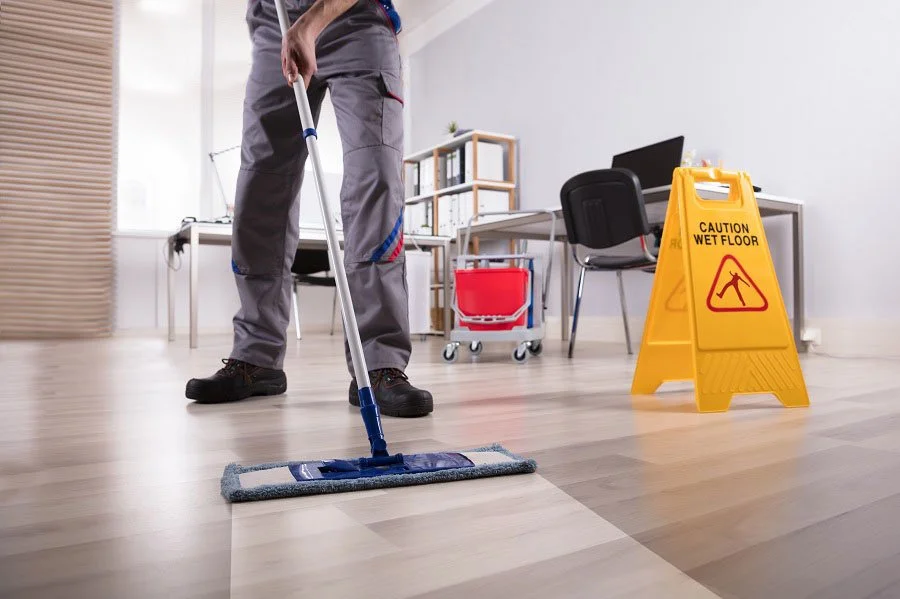 A person mopping a light wood floor in an office with a caution wet floor sign.