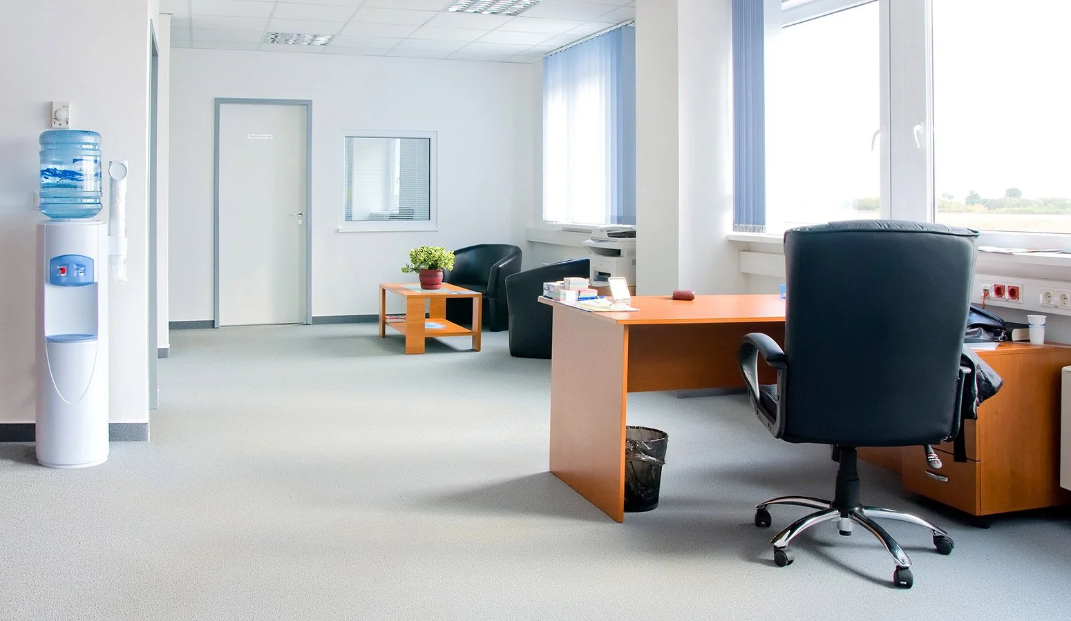 Empty office with a wooden desk, black swivel chair, small side table with a potted plant, water cooler, and large windows with blinds.