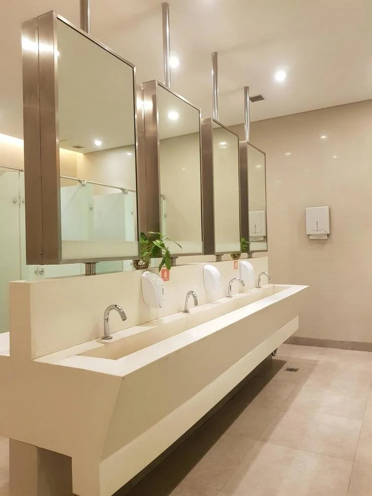 A modern public restroom with four sinks, mirrors, soap dispensers, and hand dryers on a beige wall, with a floor drain visible.