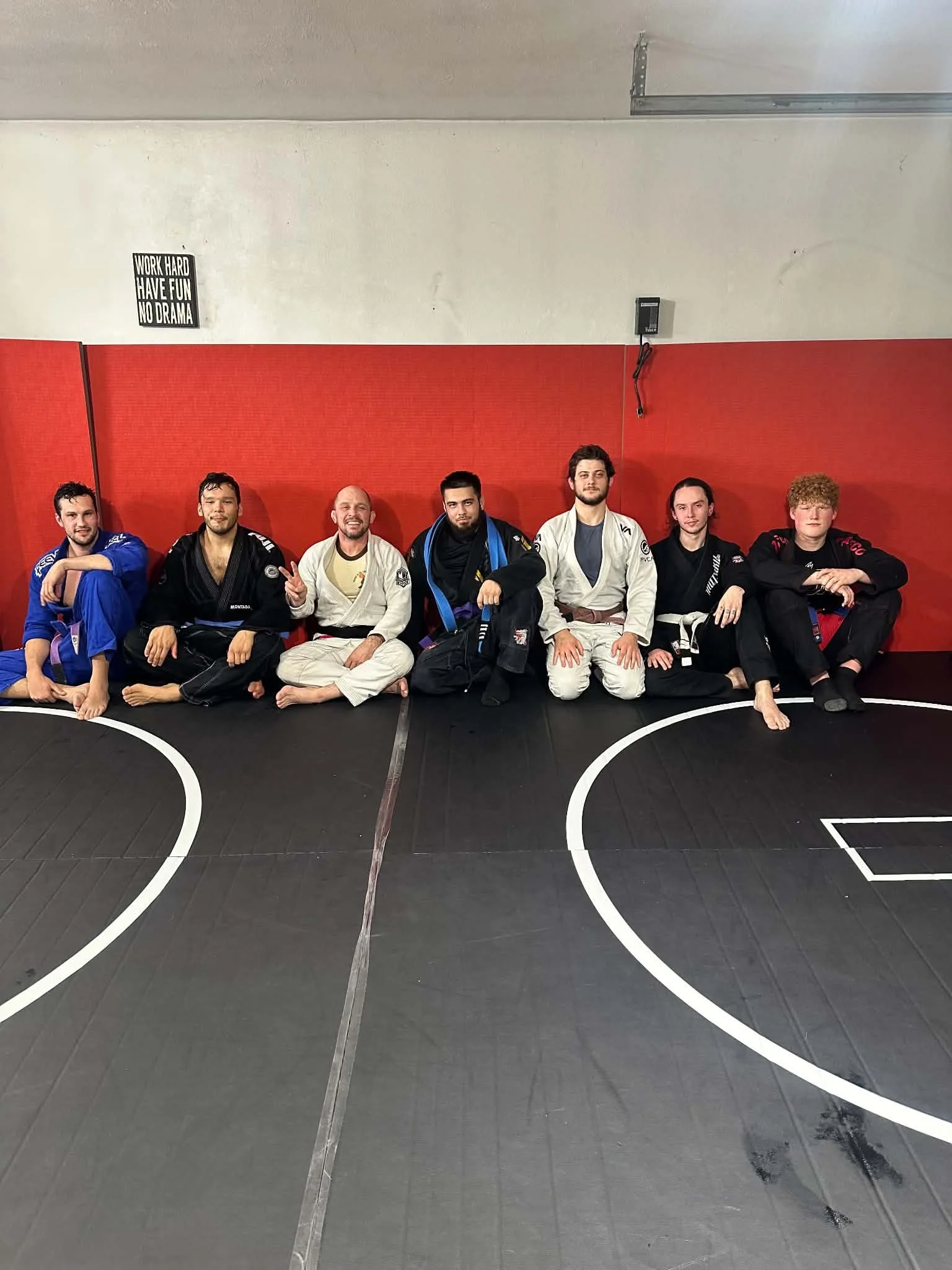 Group of seven men in Brazilian Jiu-Jitsu uniforms sitting on the mat in a martial arts gym. The red wall with a small sign and black mat flooring are visible.