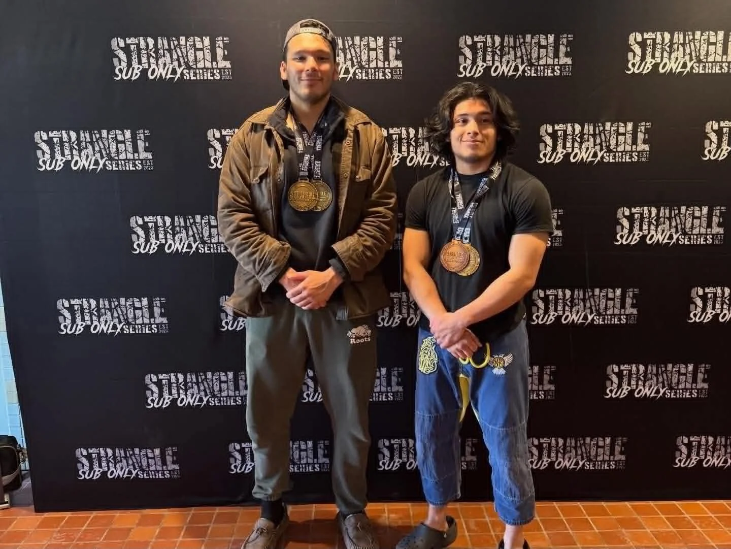Two men standing in front of a black backdrop with the words "Strangle Sub Only Series" repeated. Both are wearing medals around their necks and casual clothing. The man on the left is wearing a brown jacket, grey pants, and a cap backward. The man on the right has shoulder-length curly hair, a black t-shirt, and loose-fitting blue pants.