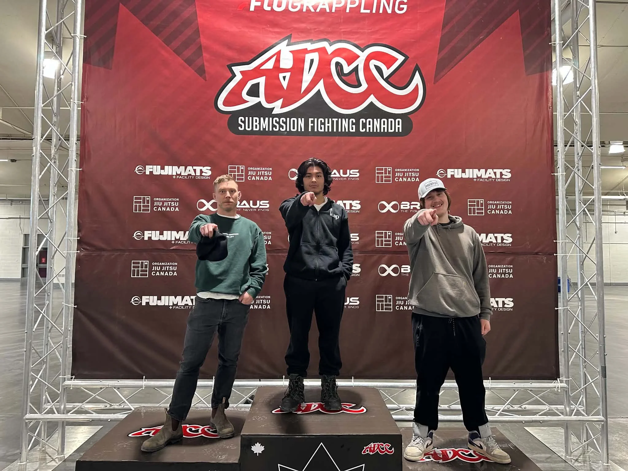 Three young men standing on a winners' podium with a red backdrop that features logos and text. The man in the center is on the top step, wearing a black hoodie, pointing at the camera. The man on the left, on the second place step, wears a green hoodie, and the man on the right, on the third place step, wears a gray hoodie and a white cap. All are posing with one hand extended forward, fists clenched.