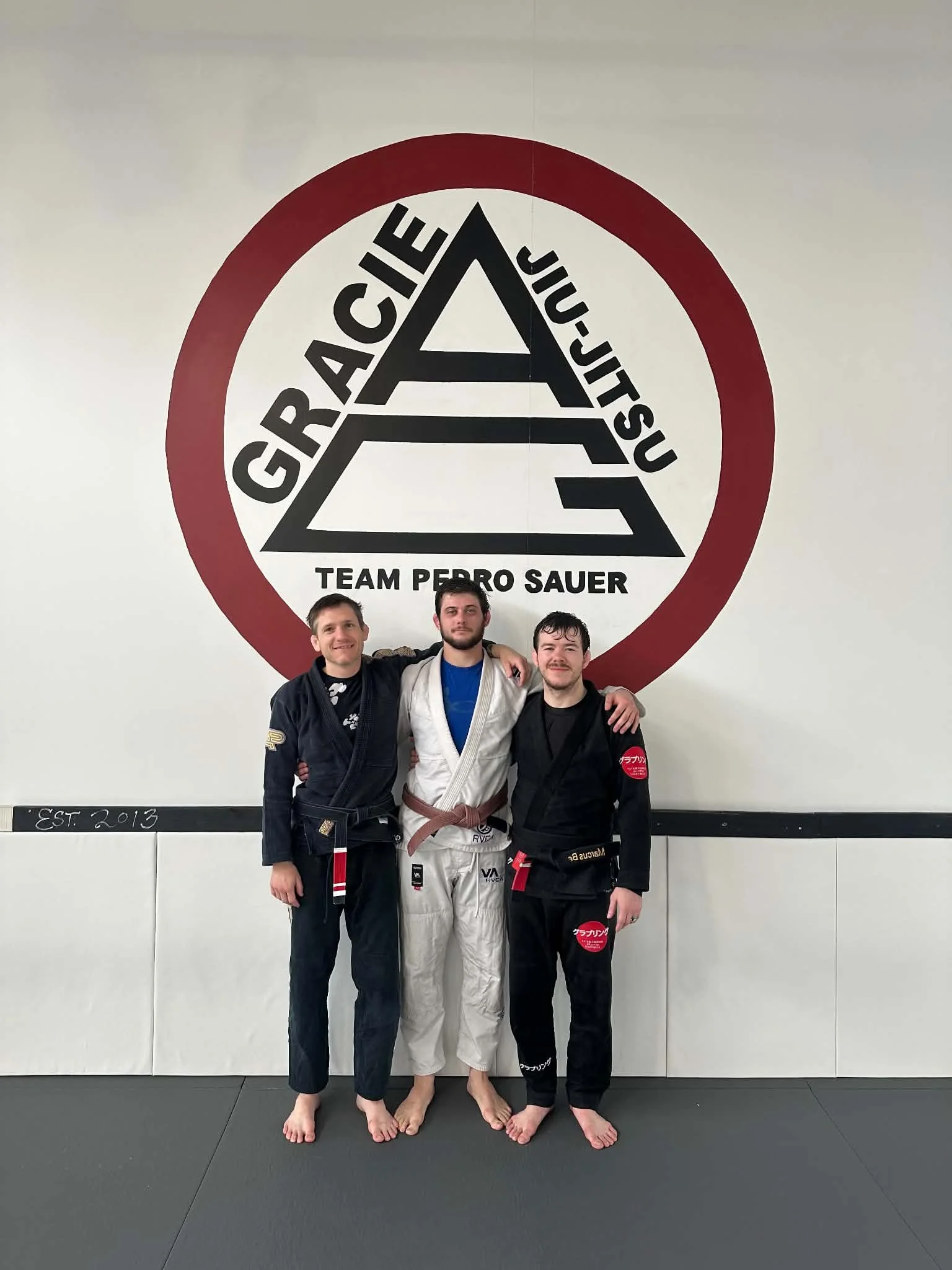 Three men standing together on a judo mat in front of a logo that reads 'GRACIE JIU JITSU TEAM PERRO SAUER' with a large stylized 'A' and a red circle around it. The men are wearing judo gis, and the middle one is in a white gi, while the others are in black gis. They are smiling and appear to be celebrating or posing after a training session or competition.