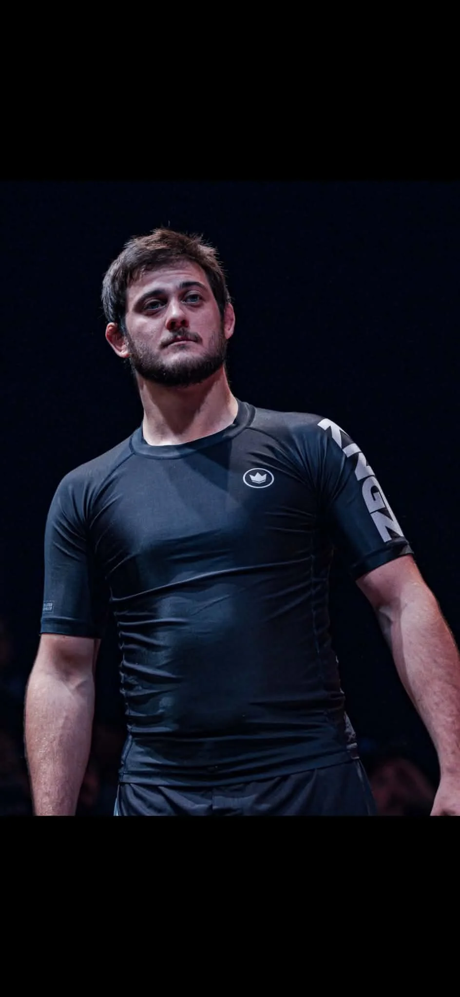 A male athlete wearing a black athletic shirt with the brand logo, standing in a dark environment, looking confident.