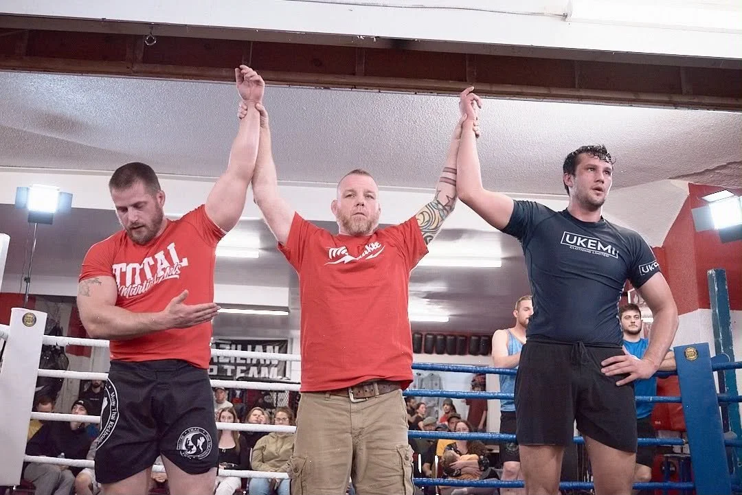 A grappler standing in a boxing ring with his arms raised by an official, one on each side, after a match. The grappler on the left wears a red shirt, and the one on the right wears a black shirt. The background shows an audience watching the event.