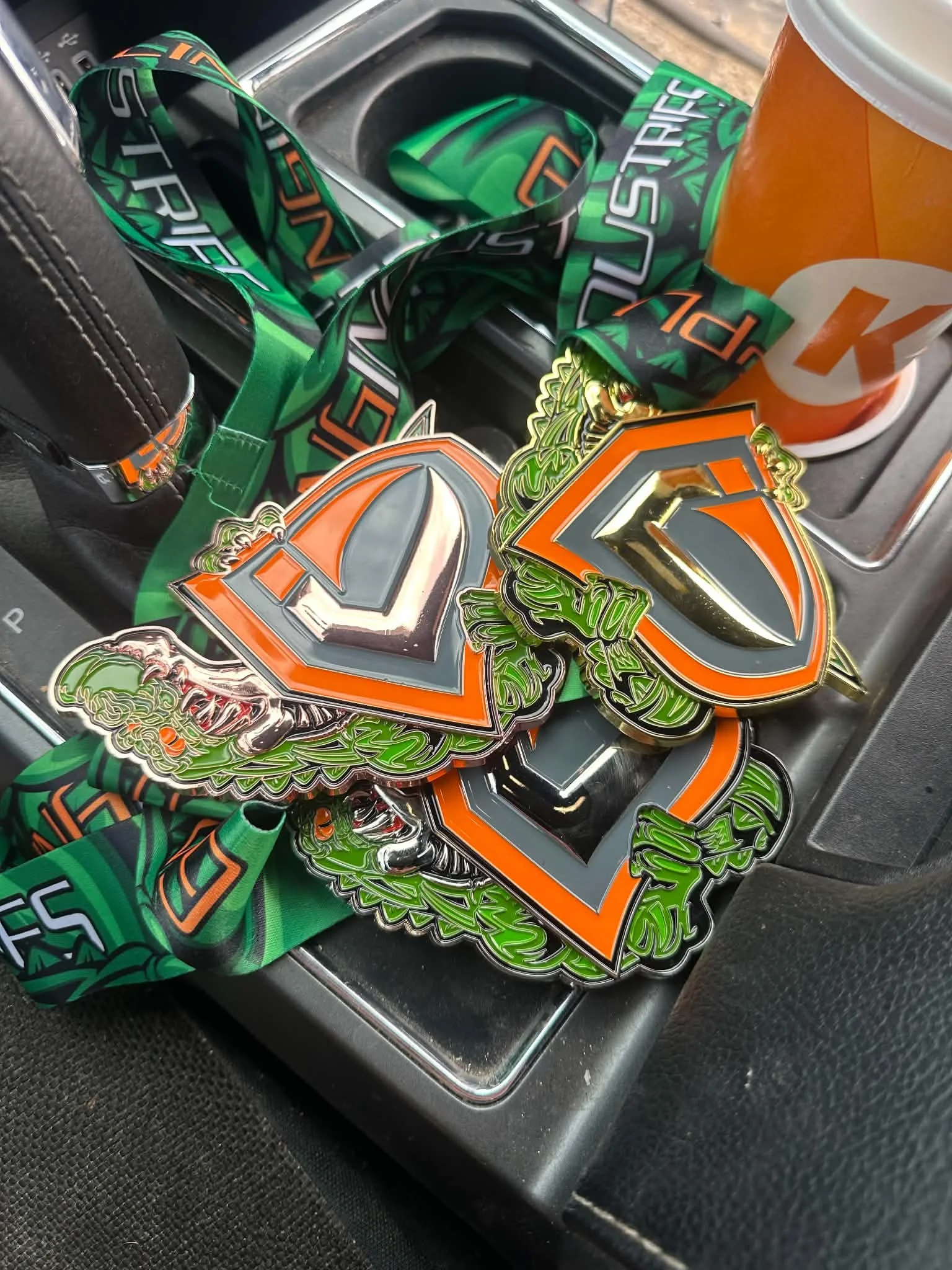 A collection of stickers, wristbands, and a cup inside a car's cup holder area. The stickers are primarily orange, green, black, and gray with bold letter designs.