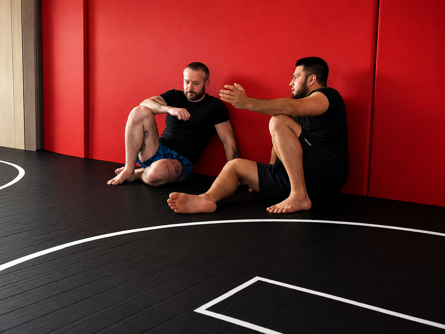Two men sitting on a black sports mat against a red padded wall, engaged in a conversation, one gesturing with his hand.