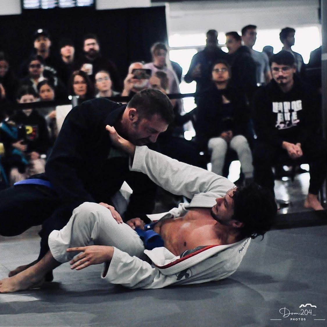 Two men practicing Brazilian Jiu-Jitsu on a mat, with one man on the ground and the other leaning over him, surrounded by a crowd of spectators.