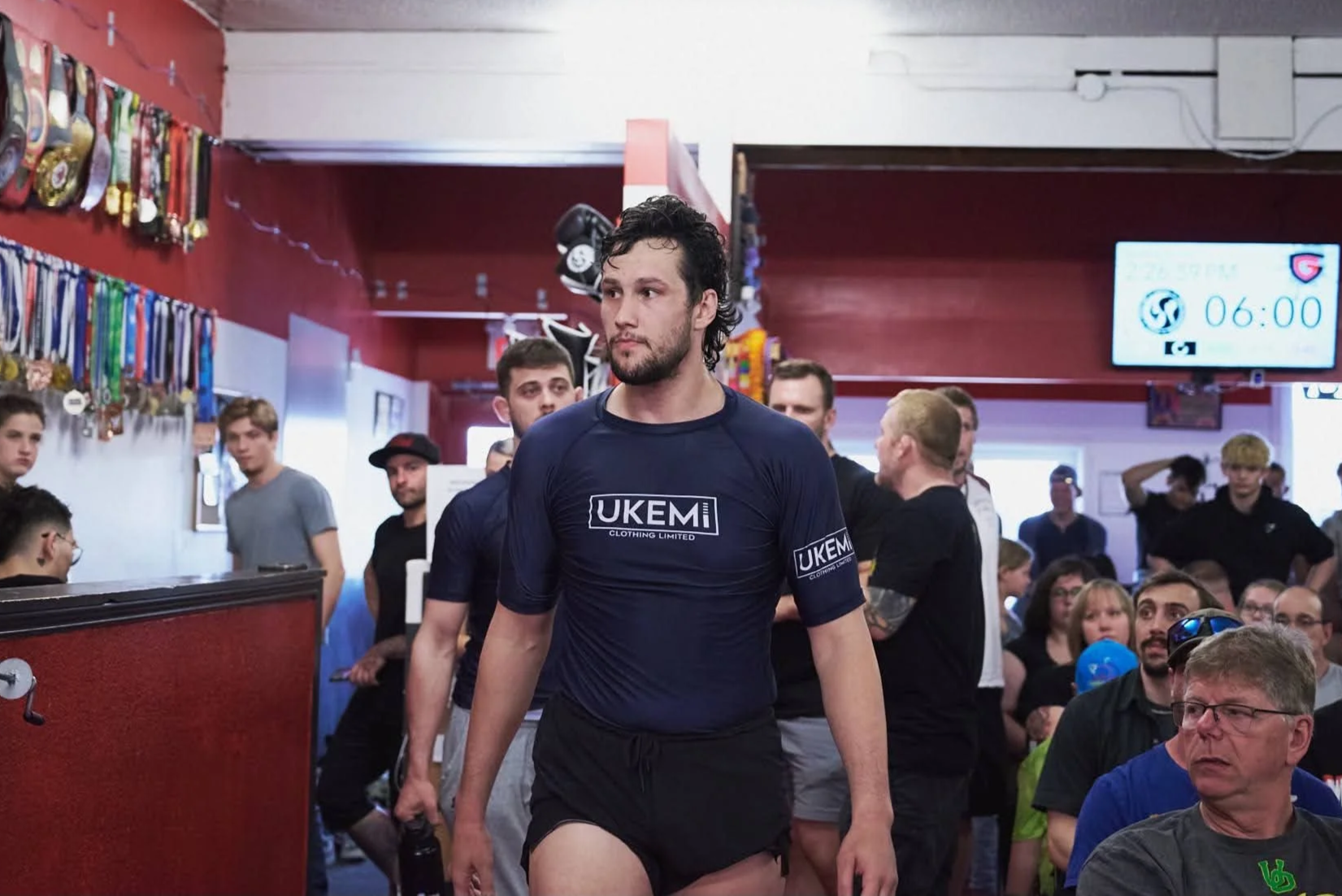A man in a navy blue T-shirt with 'UKEM Clothing Limited' on it is walking through a crowd inside a gym or event space with medals hanging on the wall and a digital screen displaying the time 06:00.