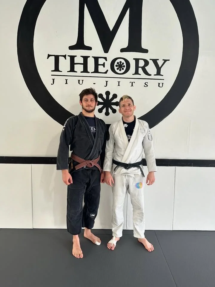 Two men in Brazilian Jiu-Jitsu gis, one in black and the other in white, standing side by side in a martial arts gym with a large 'M Theory Jiu-Jitsu' logo on the wall behind them.