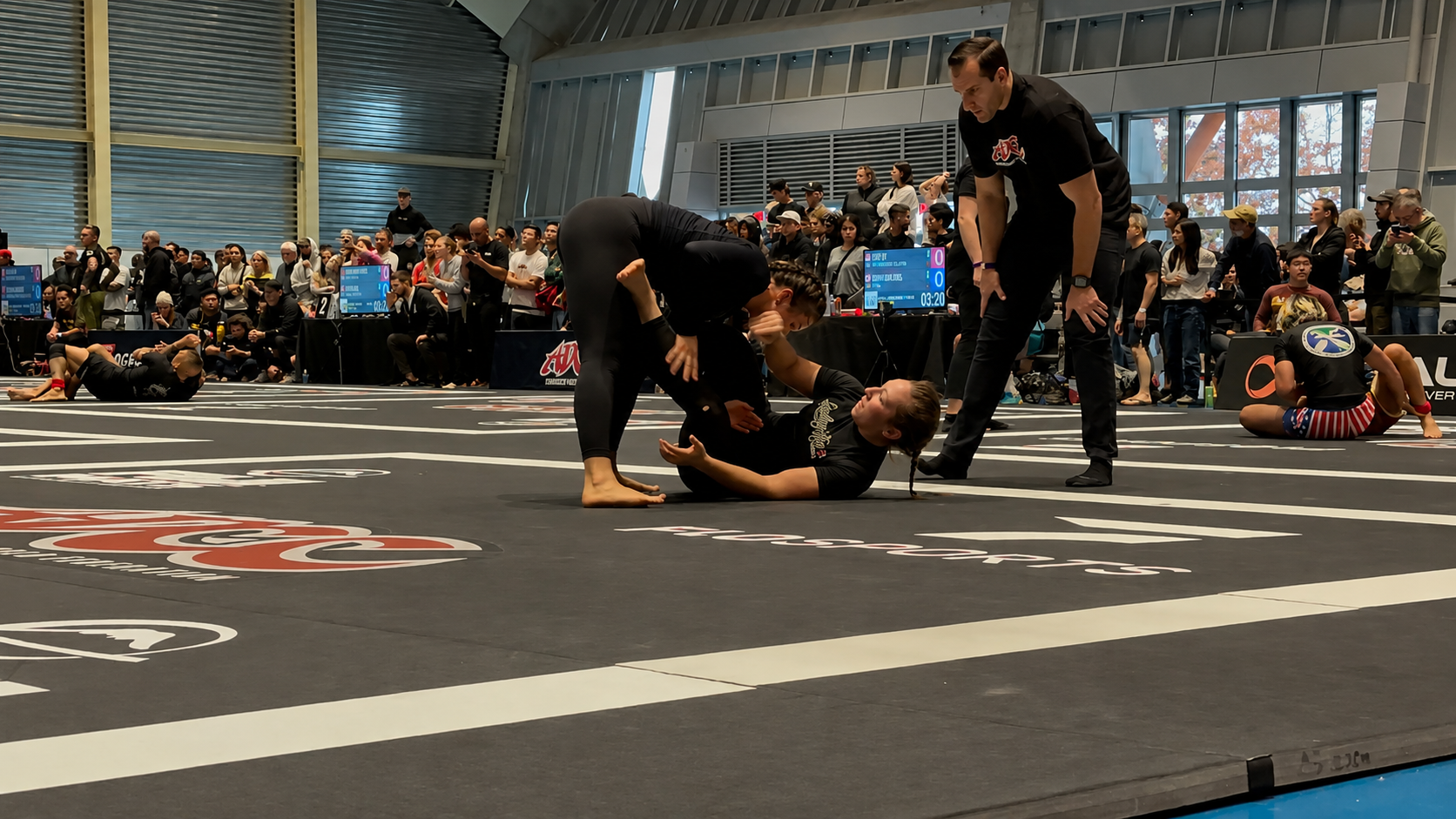 Mixed martial arts competition with female fighters on a black mat, one on her back and another over her, in front of a large crowd in an indoor arena.