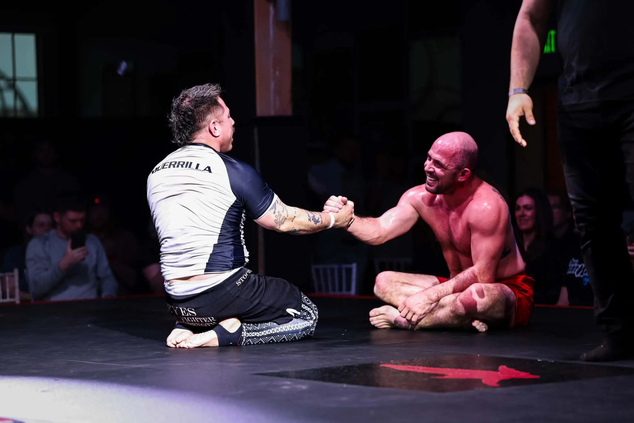Two men engaged in a Brazilian jiu-jitsu match, one kneeling and the other sitting on the mat, smiling and holding hands, with a crowd watching in the background.
