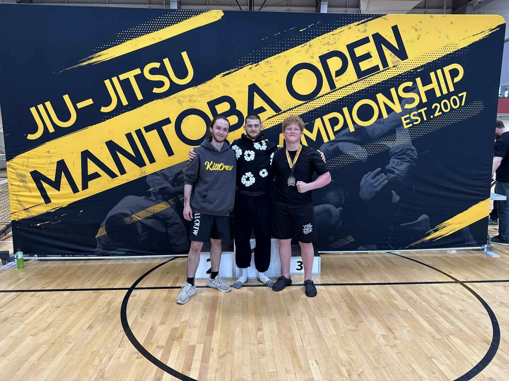 Three young male athletes standing together after a martial arts competition, pose with their arms around each other in front of a large banner that reads 'Jiu-Jitsu Open Championships Est. 2007.' They are wearing casual athletic clothing and standing on a podium marked with the number 3. The setting is indoors on a wooden gymnasium floor.
