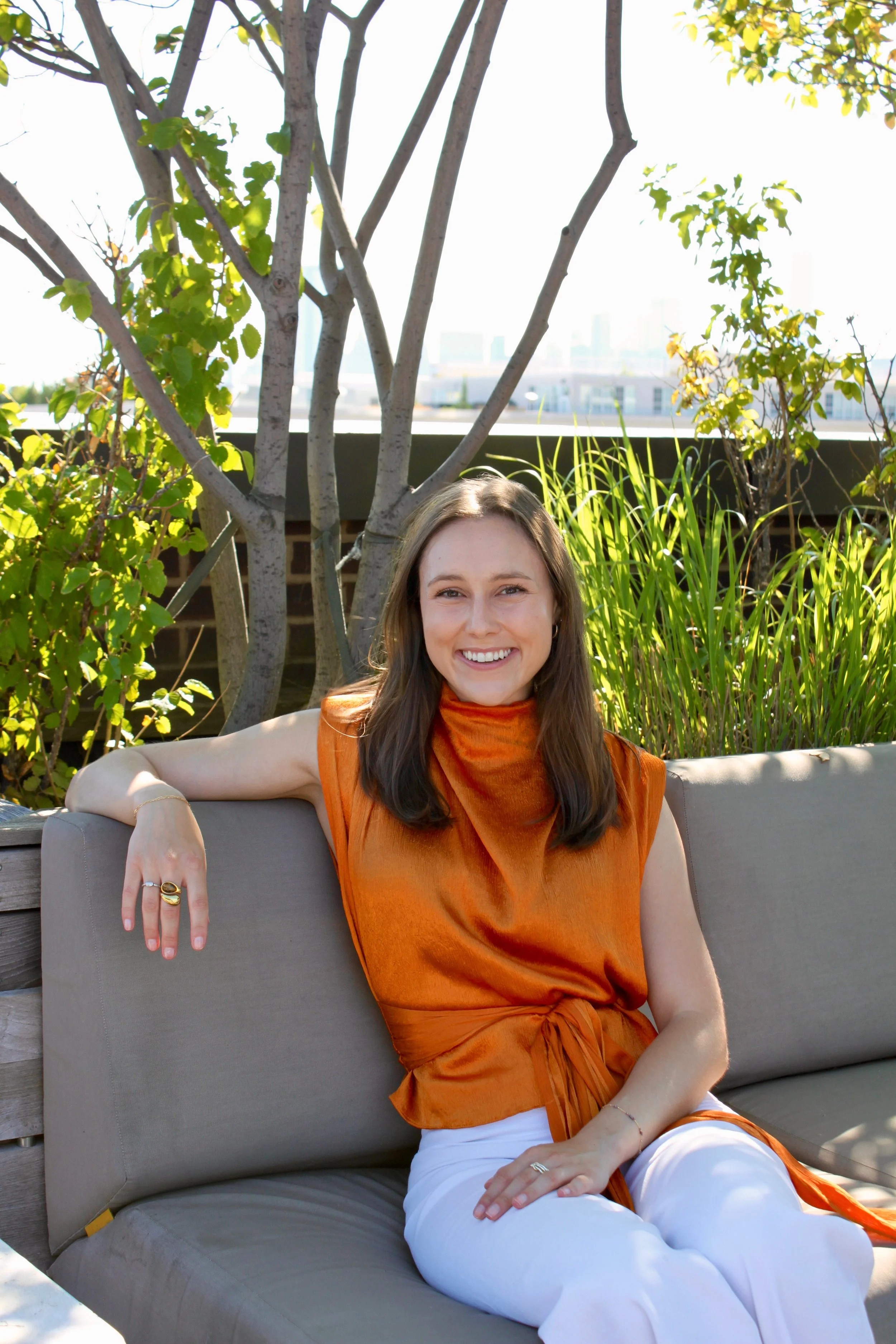 A smiling woman with long brown hair, wearing an orange sleeveless top and white pants, sits on a cushioned outdoor lounge chair surrounded by green plants and trees, with city buildings visible in the background under bright sunlight.