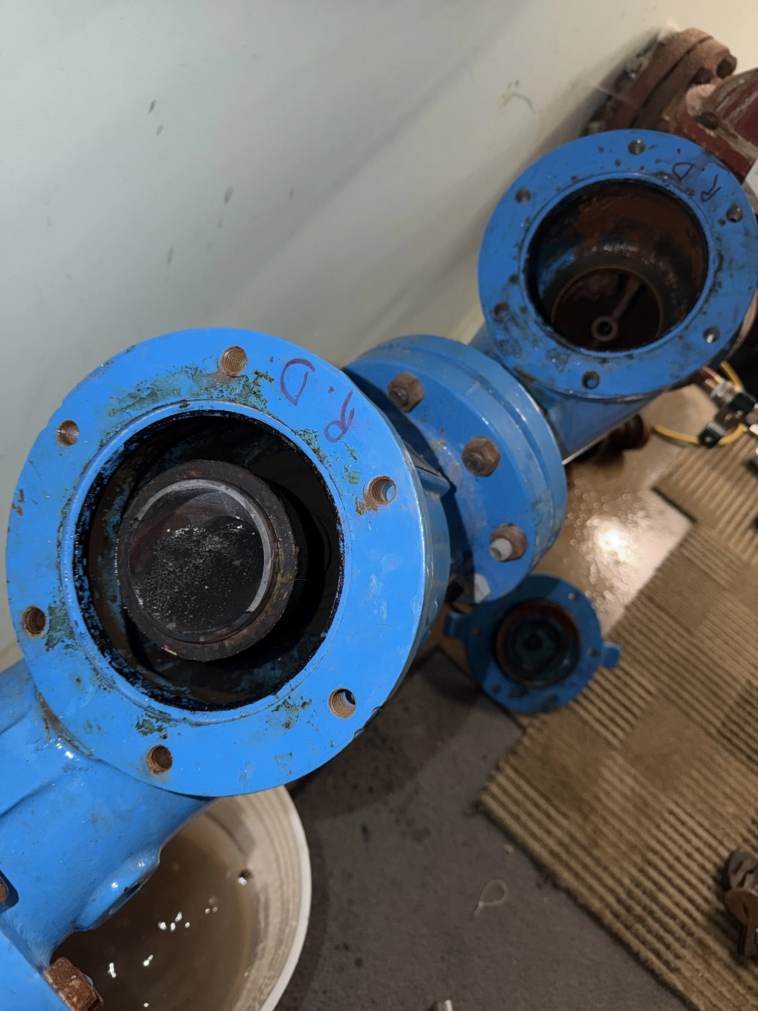 Blue industrial pipes with flanged ends, some rusty and with a few markings, arranged in a row on a concrete floor. There are buckets and a mat nearby.