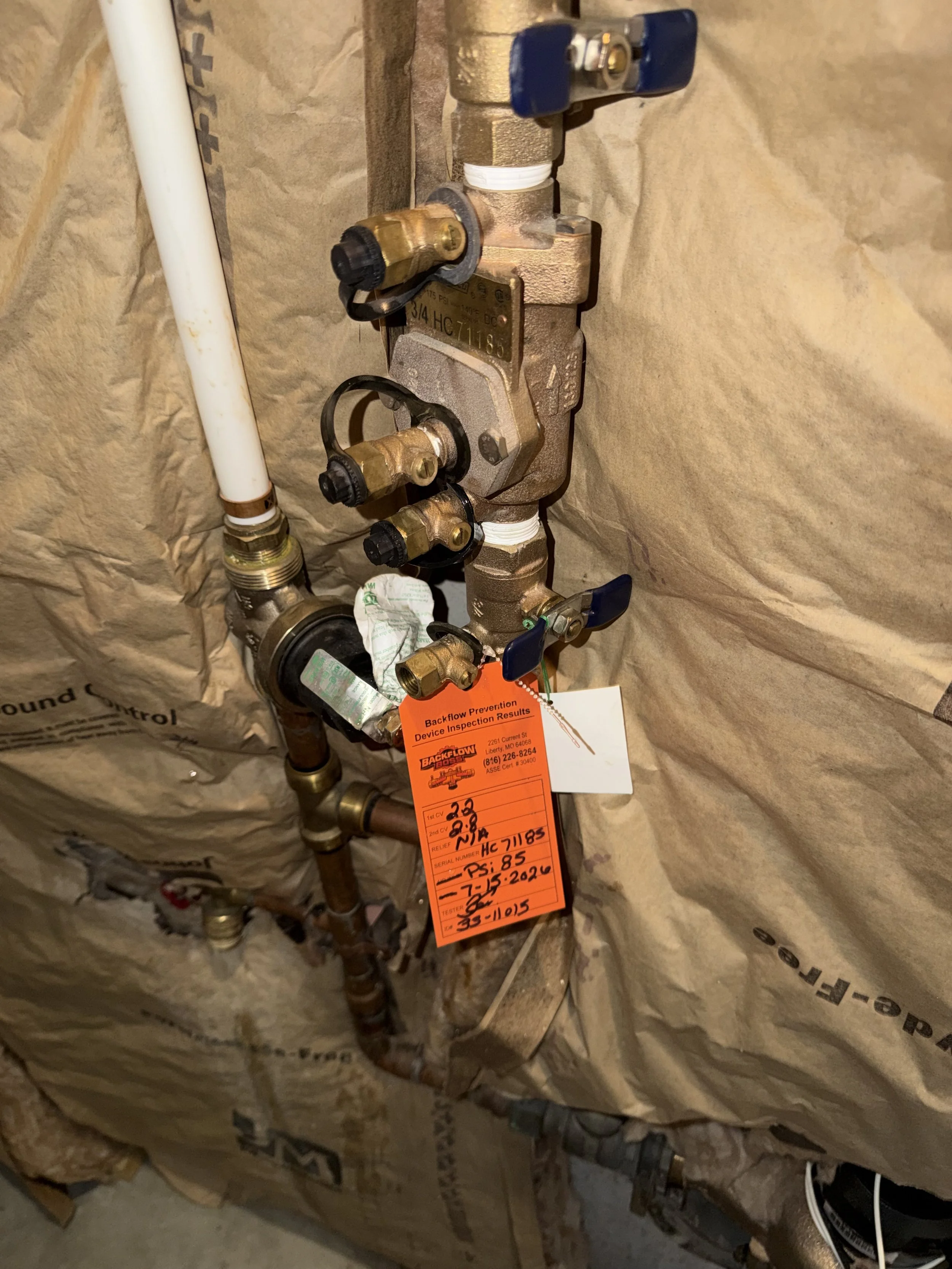 A plumbing backflow prevention device installed on a pipe system, with orange inspection tags hanging from it, surrounded by brown insulation material.
