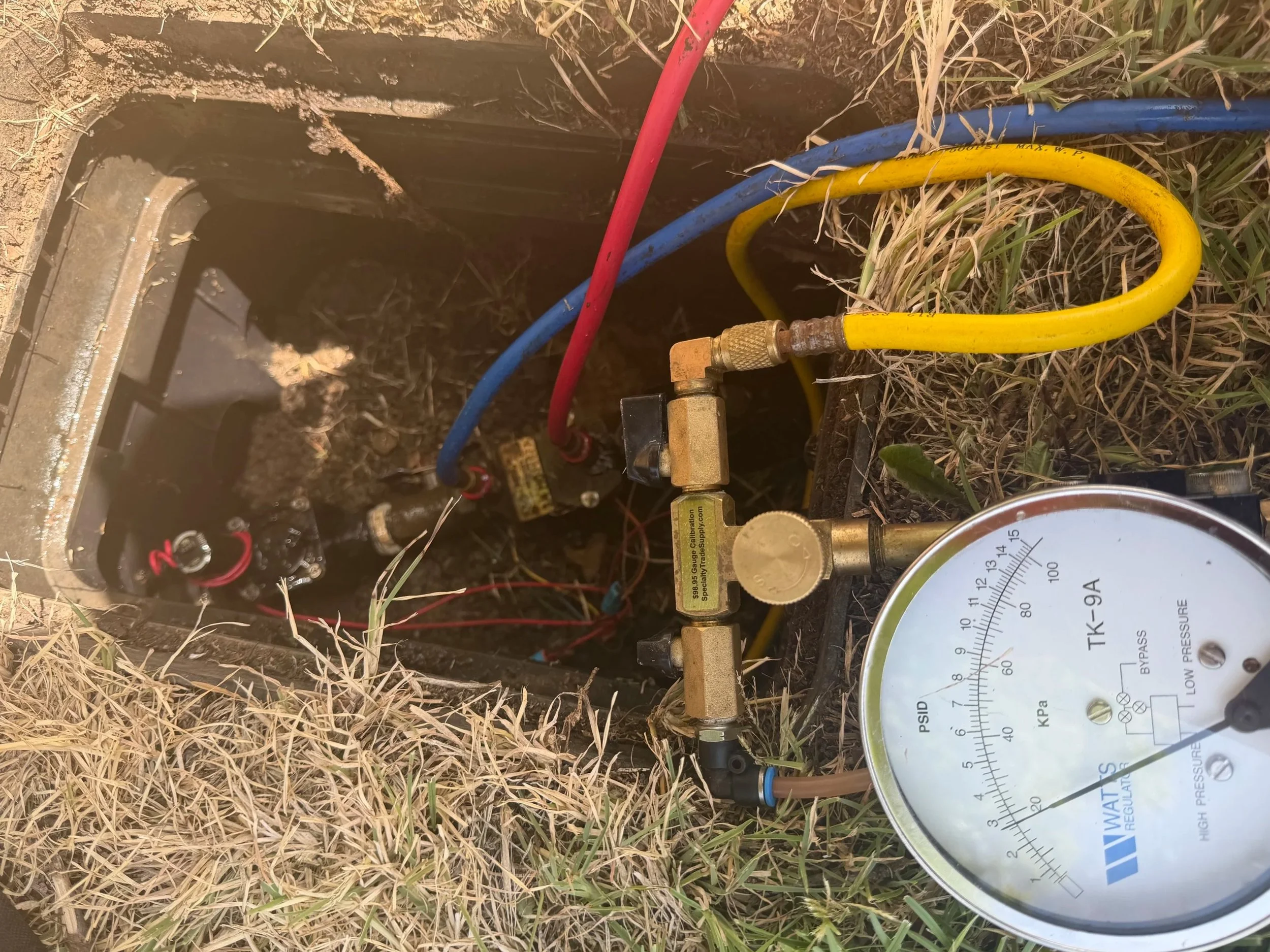 Close-up of a gauge measuring water pressure with multiple colorful hoses connected, situated outdoors in a grassy area.