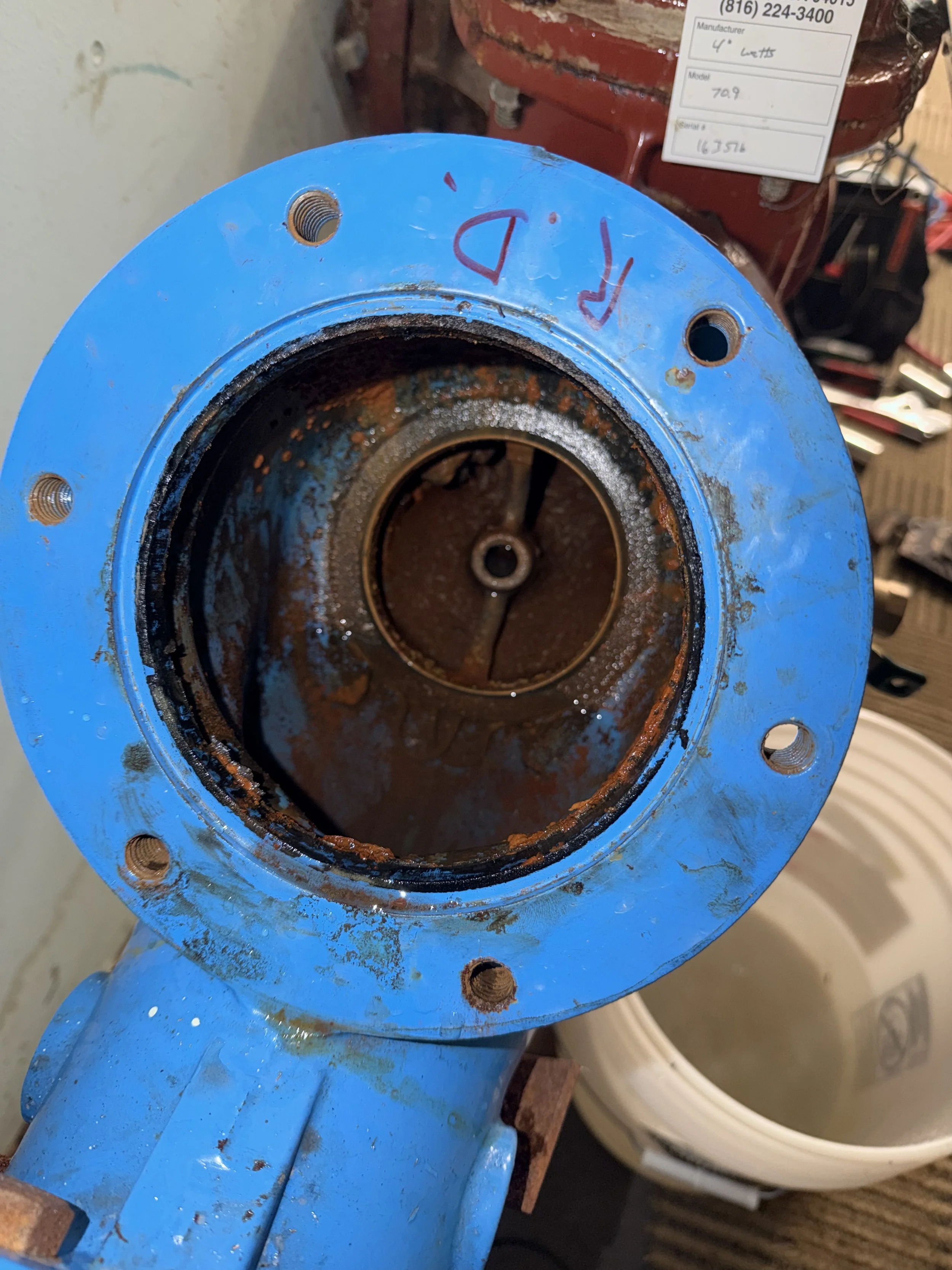 Close-up of a rusty blue pipe flange with four bolt holes, showing signs of corrosion and dirt inside, and a red machinery part with a label in the background.