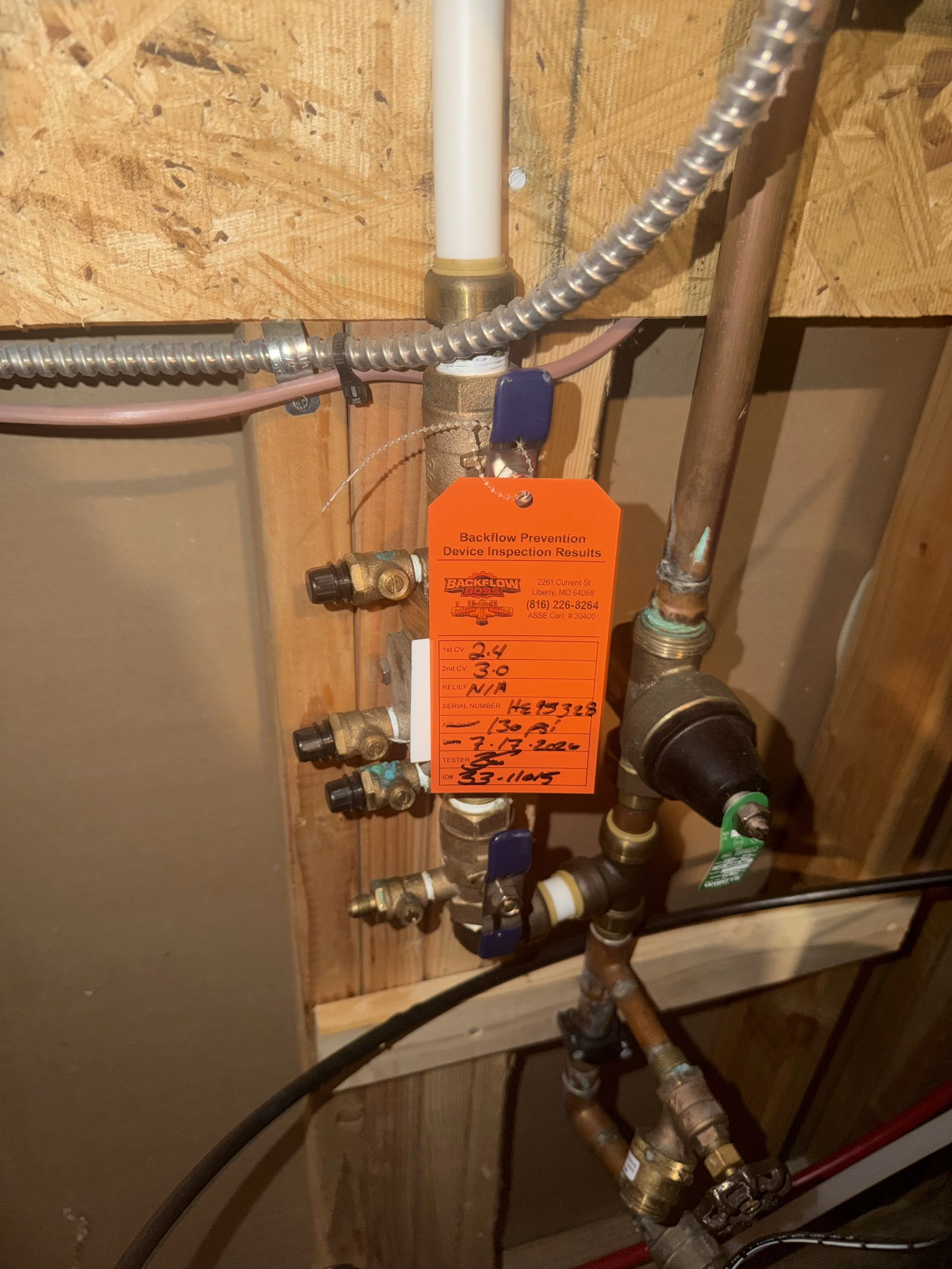 A plumbing assembly with pipes, valves, and a backflow prevention device attached to a wooden wall, with a bright orange inspection tag.