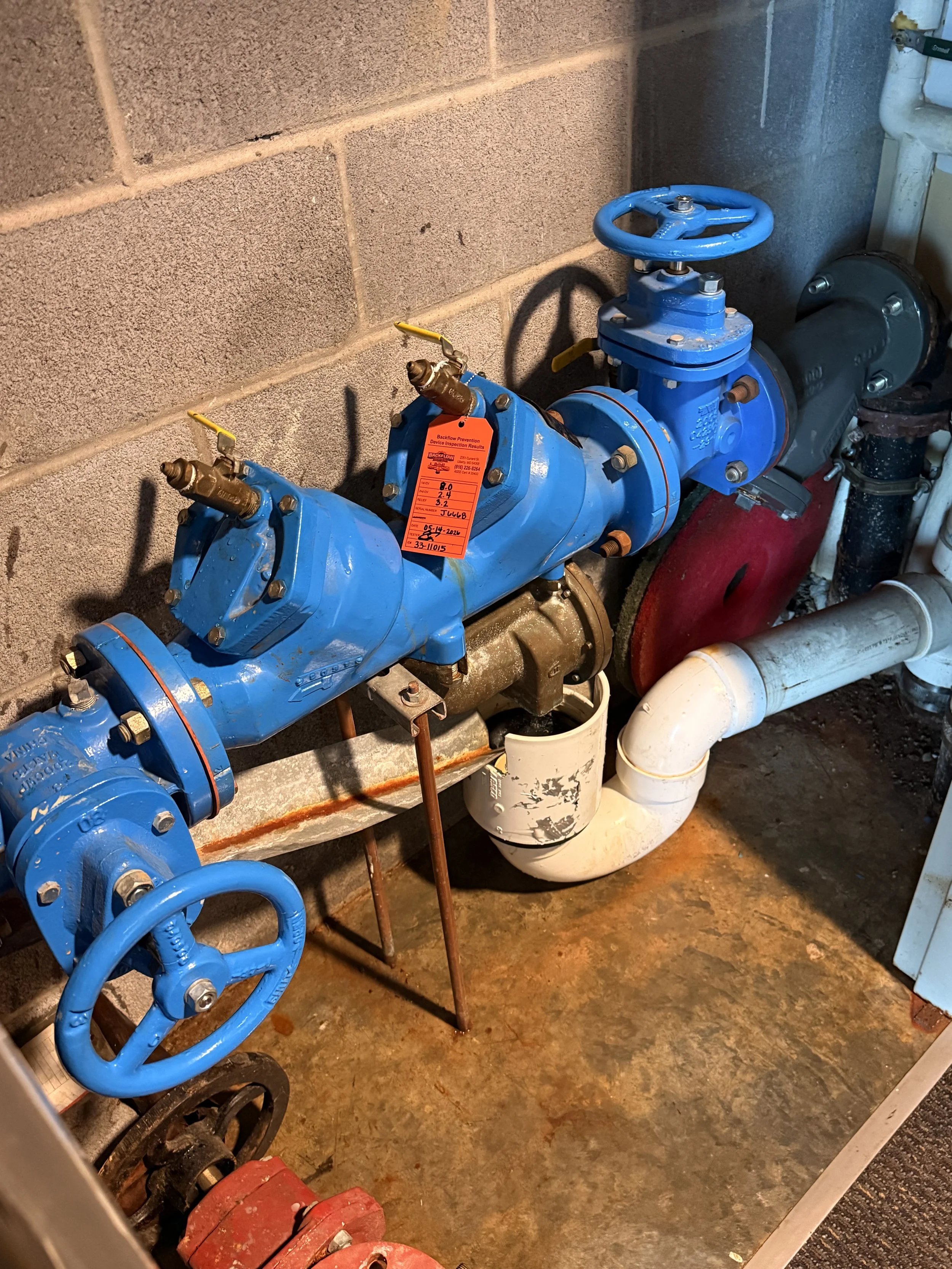 A blue water valve connected to a pipe system in a utility room or basement with exposed brick wall and concrete floor.