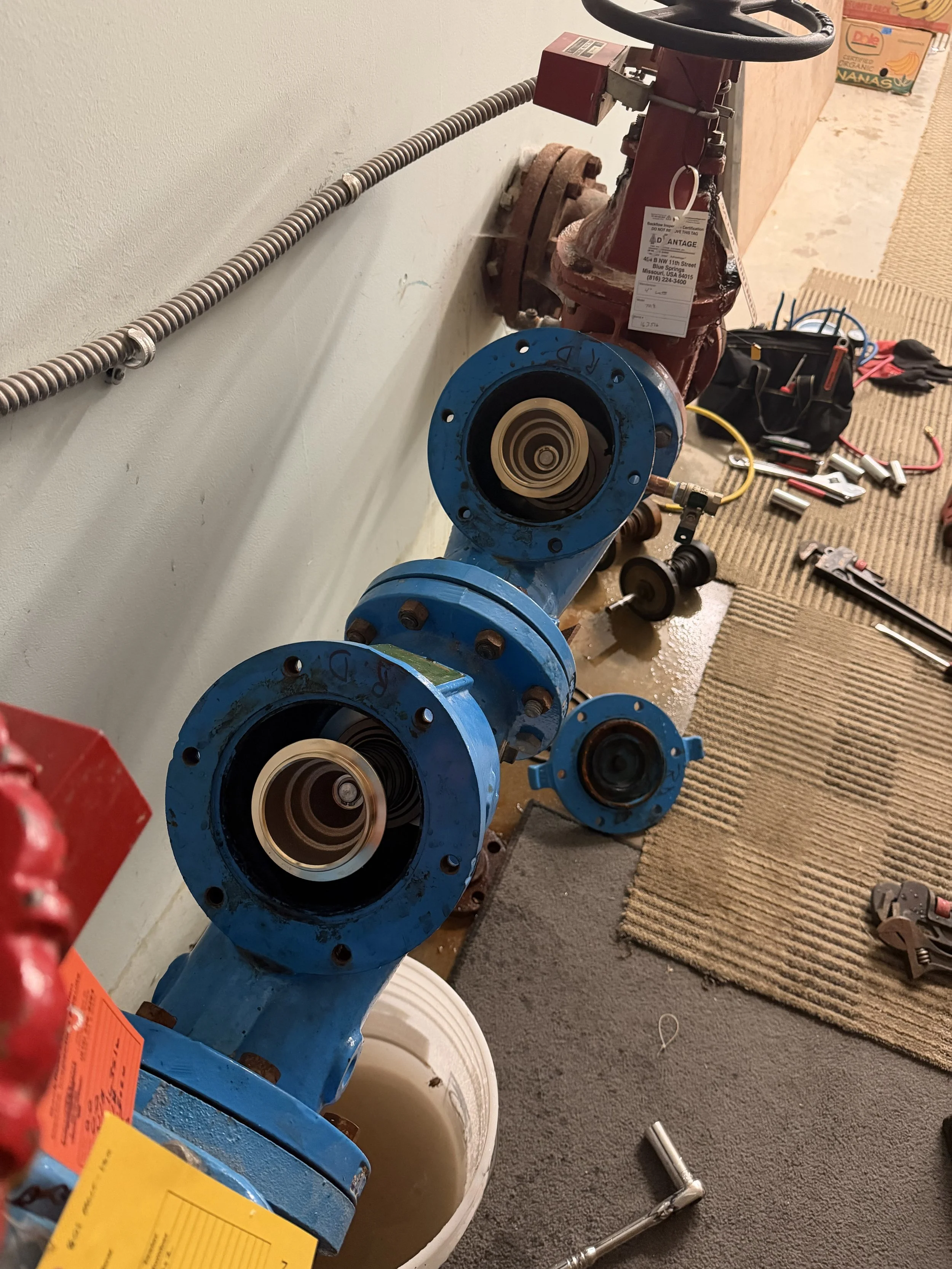 A finished section of a plumbing system with blue valve housings, an exposed pipe connection, and various tools on the floor in what appears to be a construction or maintenance setting.