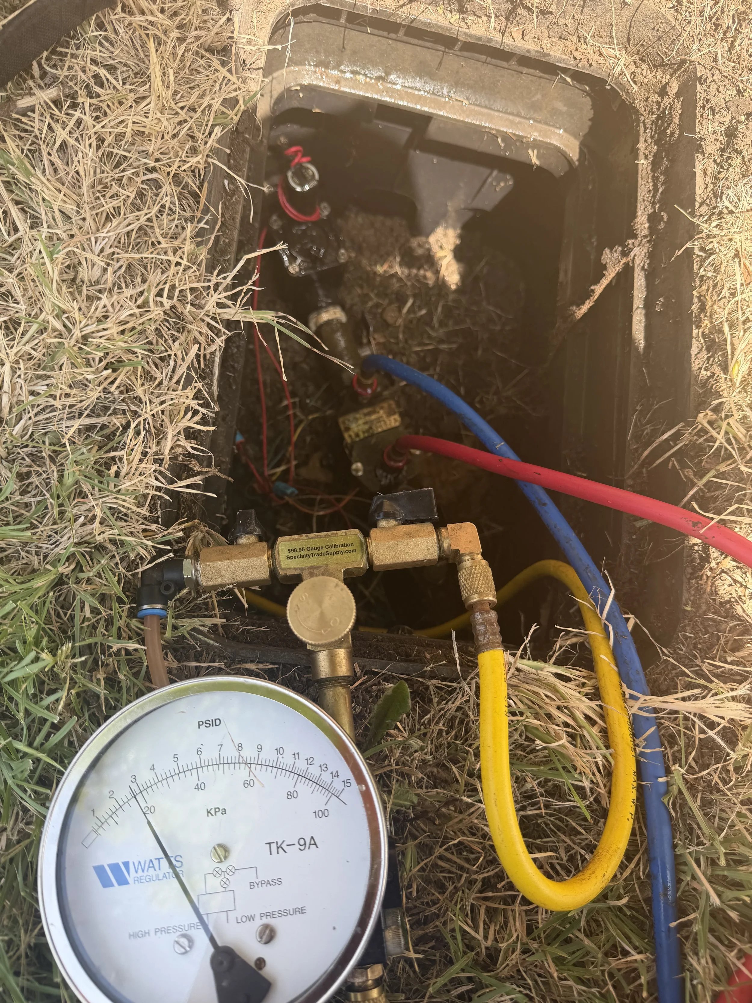 Pressure gauge connected to pipes, with yellow, red, and blue hoses, surrounded by grass and soil.