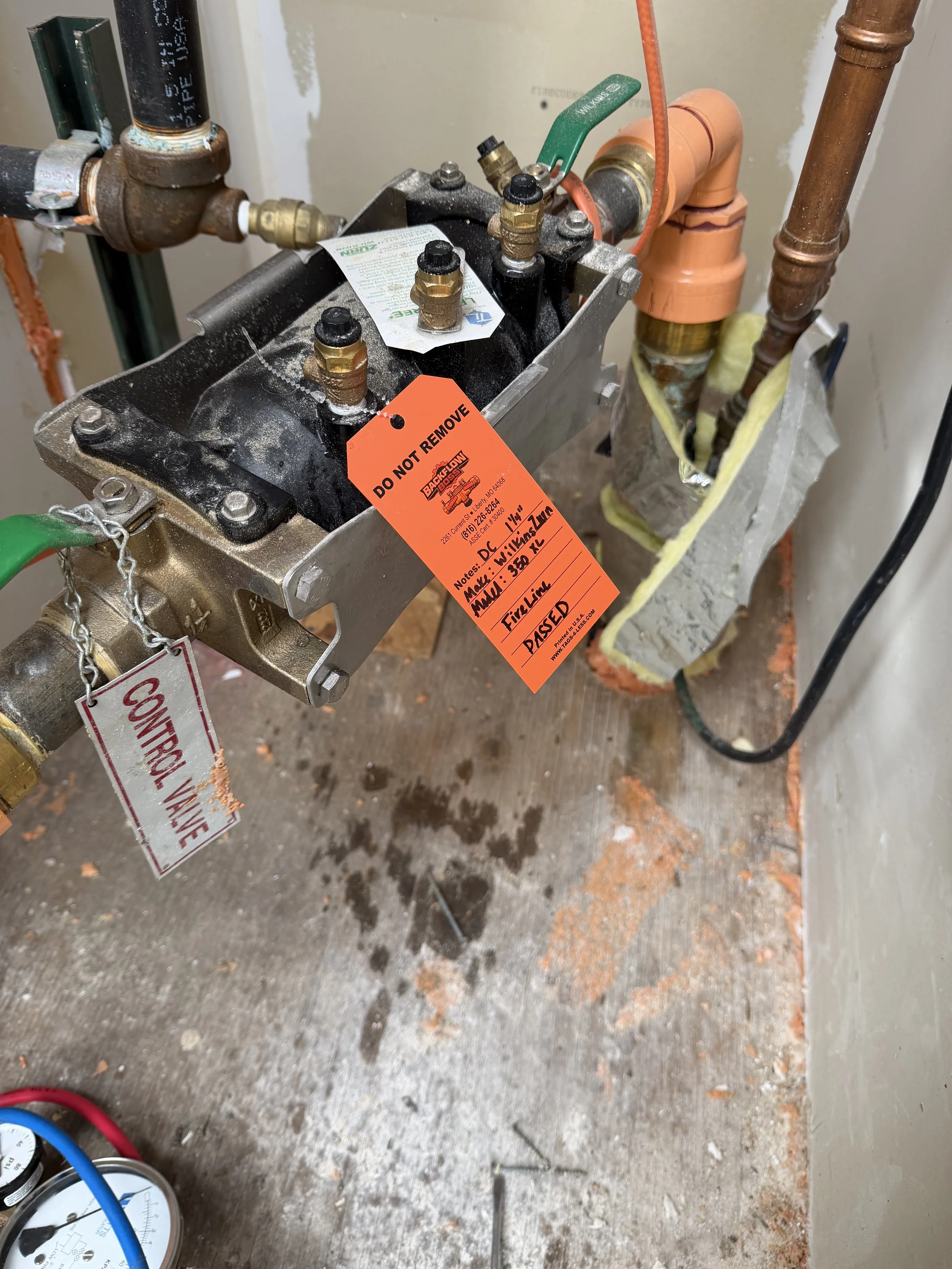Close-up of plumbing work with pipes, valves, and a control box with orange tags, on a construction site with a concrete floor.