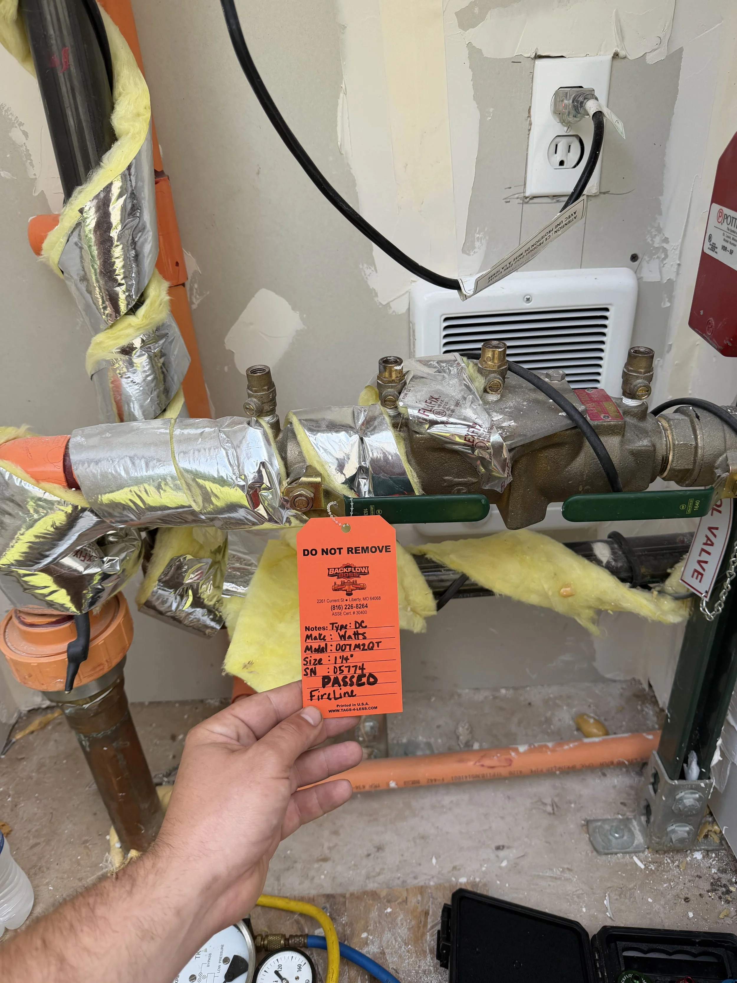 Close-up of a hand holding an orange tag labeled 'PASSED' in front of plumbing and electrical components, including pipes wrapped in reflective insulation, a wall outlet, and a fire alarm. The area appears to be under construction or repair.