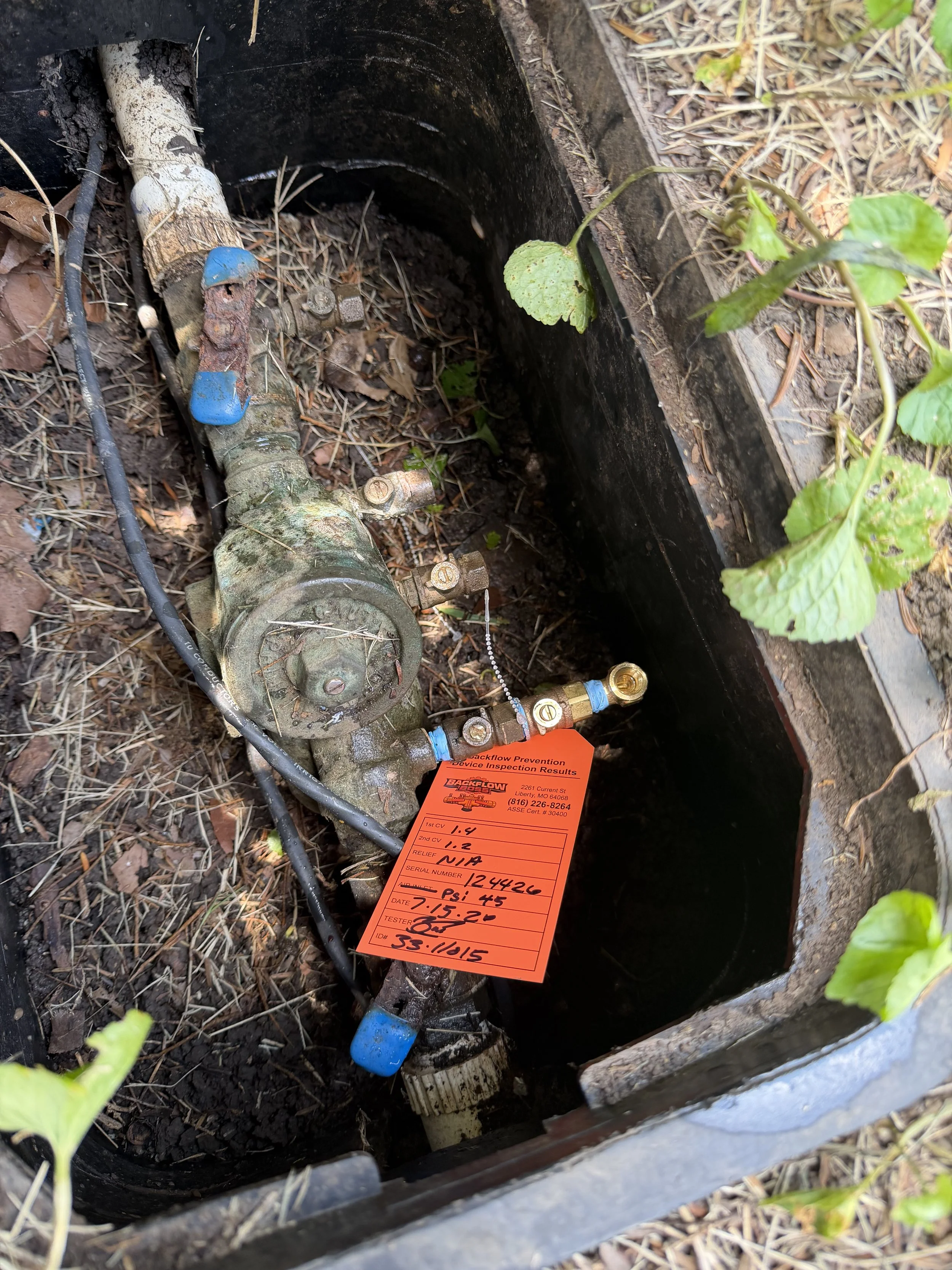 Close-up of a plumbing valve and pipes with an orange inspection tag attached, situated in an outdoor ground area with dry leaves and plants surrounding it.
