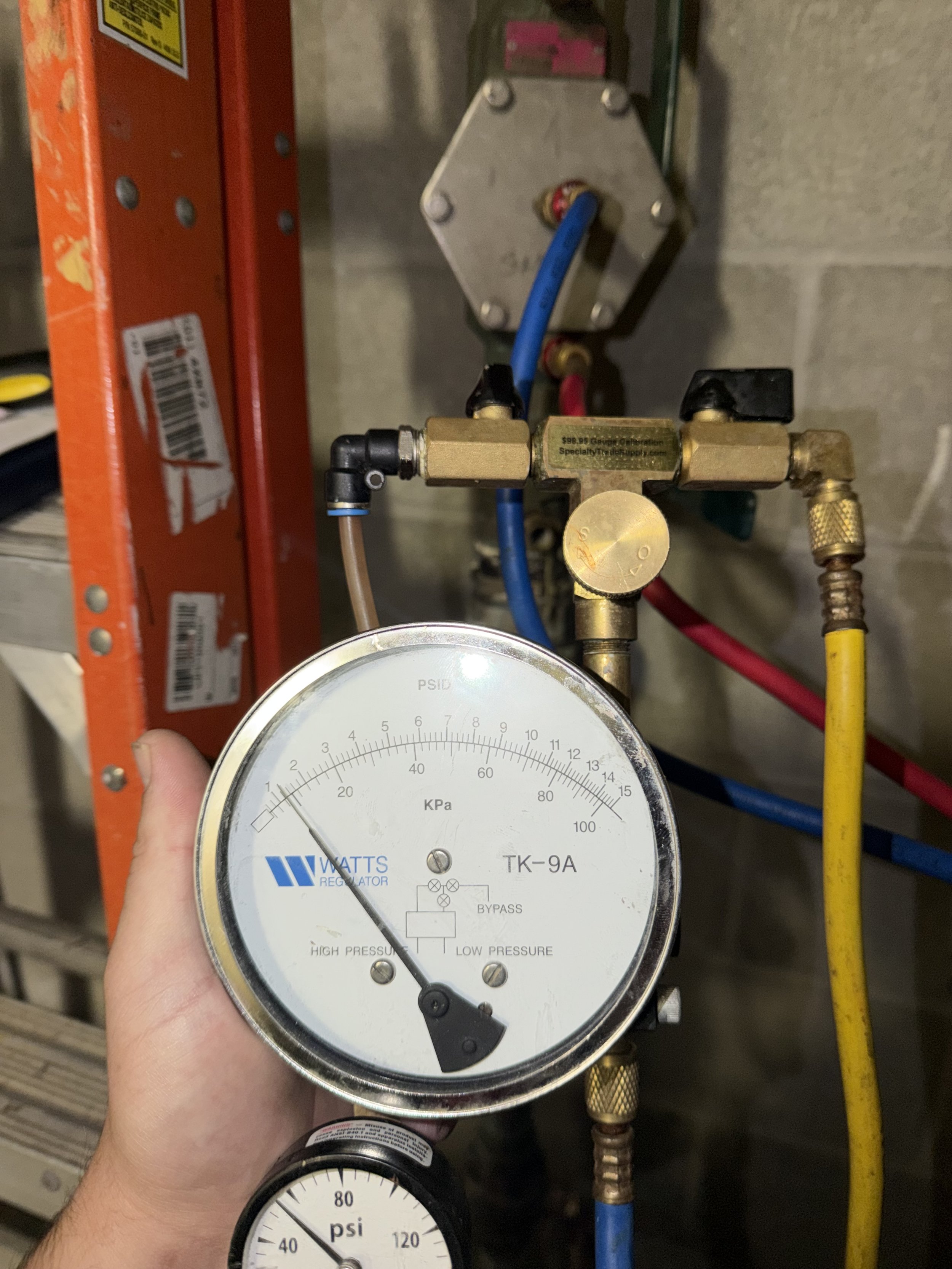 A person holding a pressure gauge with black needle indicating pressure in kilopascals, connected to a series of pipes and valves in a mechanical or HVAC setting, with a storage rack and wall in the background.