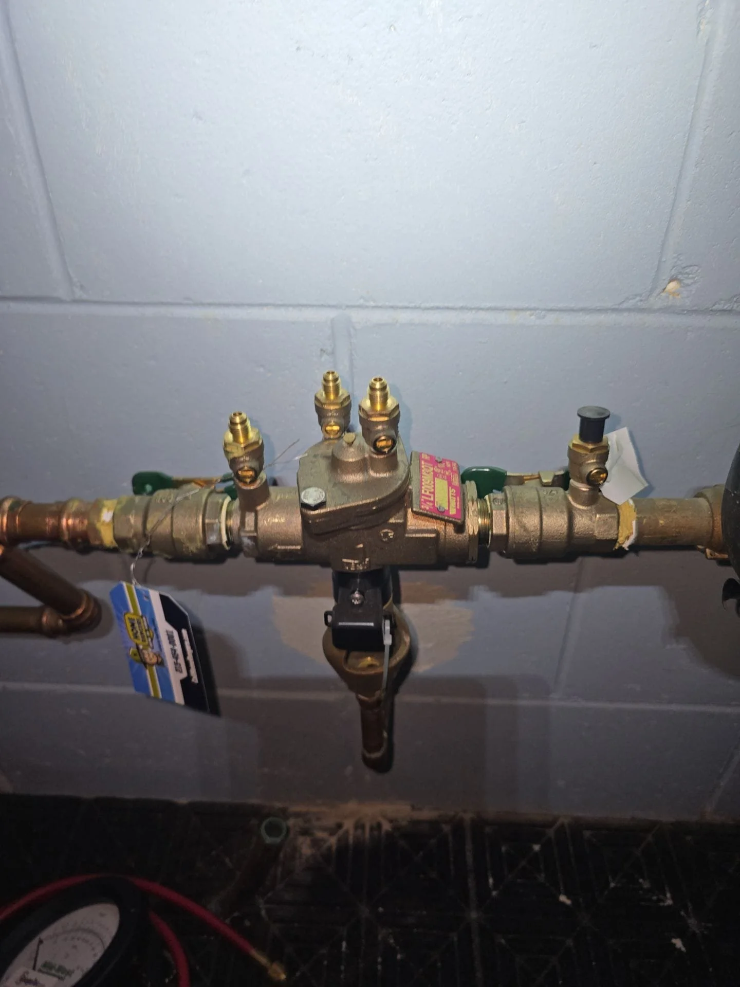 A water shutoff valve with brass fittings mounted on a wall, connected to copper pipes, with a black lever handle and a pink tag.