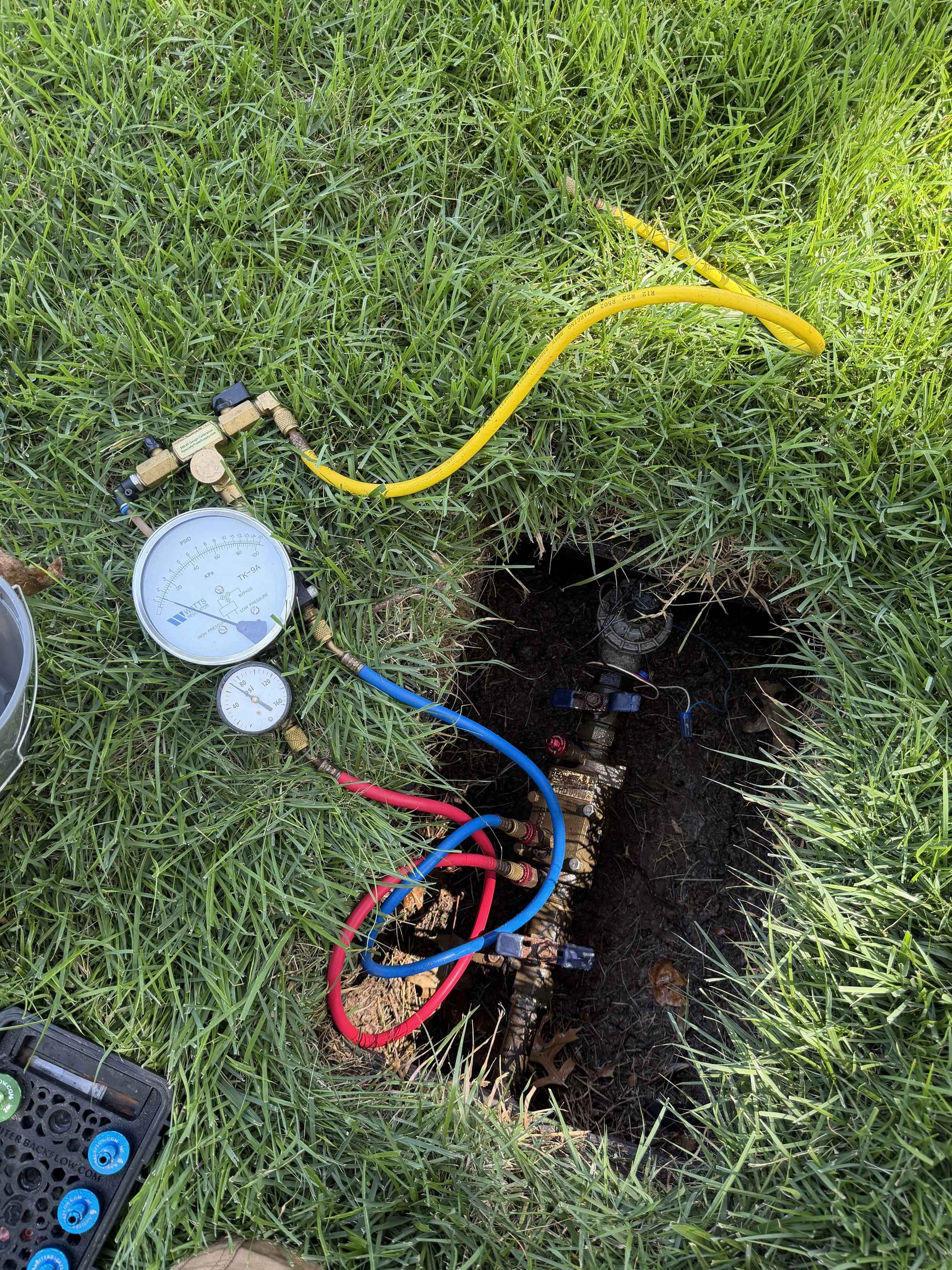 An underground utility inspection with various equipment, including gauges, hoses, and electrical connections, surrounded by green grass, partially dug trench, and a tool case.